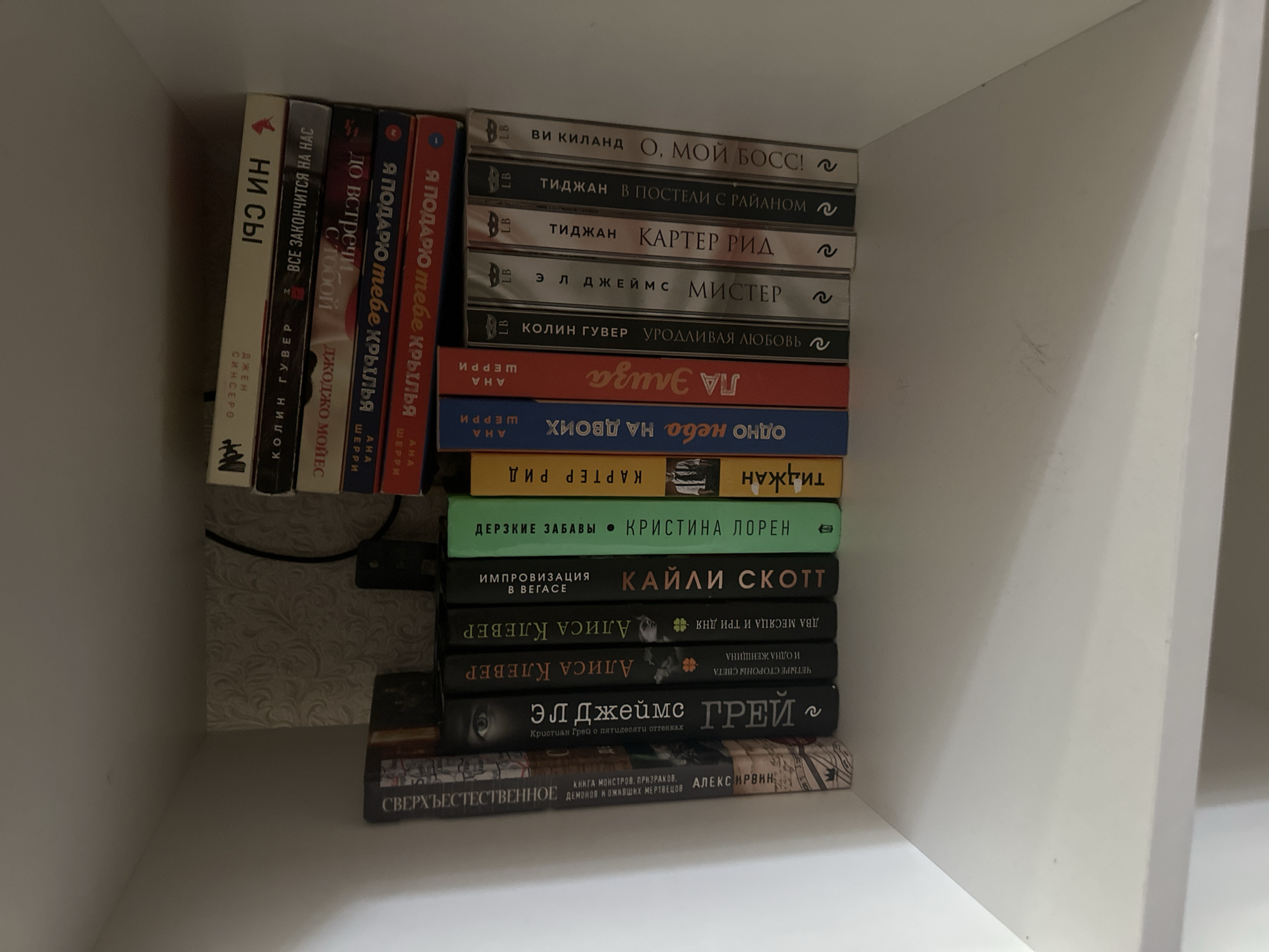 The image shows a bookshelf filled with books. The books are arranged in stacks and rows. Some of the books are standing upright, while others are lying down. The bookshelf is made of white material and is positioned in a well-lit room. The background is a light gray, textured wallpaper. The specific location in the city of Vologda, Russia, cannot be determined from the image alone.