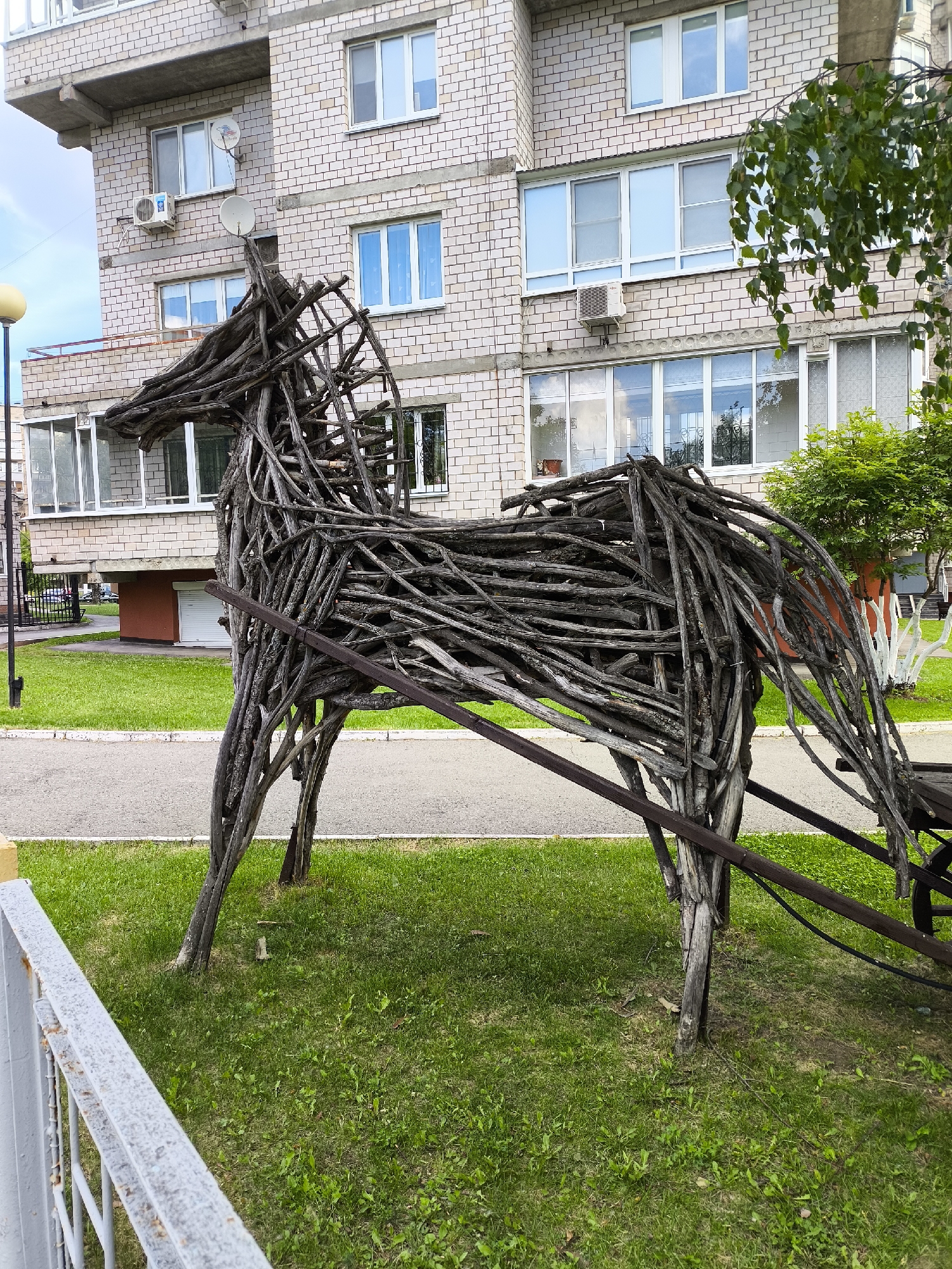 This image captures an outdoor urban scene in Izhevsk, Russia. The central and dominant subject is a life-sized sculpture of a horse, constructed entirely from numerous interlaced and bound tree branches and twigs. The sculpture is dark grey-brown, indicating weathered wood, and depicts the horse in profile, facing left, with its head slightly lowered. A dark brown wooden shaft extends backward from the horse's mid-section, suggesting it is part of a harness for an unseen carriage or sleigh, a segment of which is visible on the far right as a dark wooden, wheel-like structure.
The sculpture rests on a patch of short, bright green grass. Immediately behind the grassy area, a light grey asphalt or concrete path, edged by a white curb, runs horizontally across the frame. In the background, a multi-story residential building, constructed with light-colored brick, rises. The building features multiple rectangular windows with white frames; many windows exhibit a light blue reflection of the sky. Security bars or decorative grates are visible behind several lower-level windows. Two white satellite dishes and at least two external air conditioning units are affixed to the building's facade. Concrete balconies or protruding architectural elements are also present on various floors.
In the bottom left foreground, a grey metal fence, exhibiting patches of rust, is partially visible. A street lamp with a yellow-domed light fixture is present on the far left. To the right of the horse sculpture, a small, vibrant green bushy tree stands, with another tree featuring a white-painted lower trunk visible behind it. A branch adorned with green leaves extends into the frame from the upper right corner. The sky above the building is bright and appears to be overcast or partly cloudy. No people or animals are present in the scene.