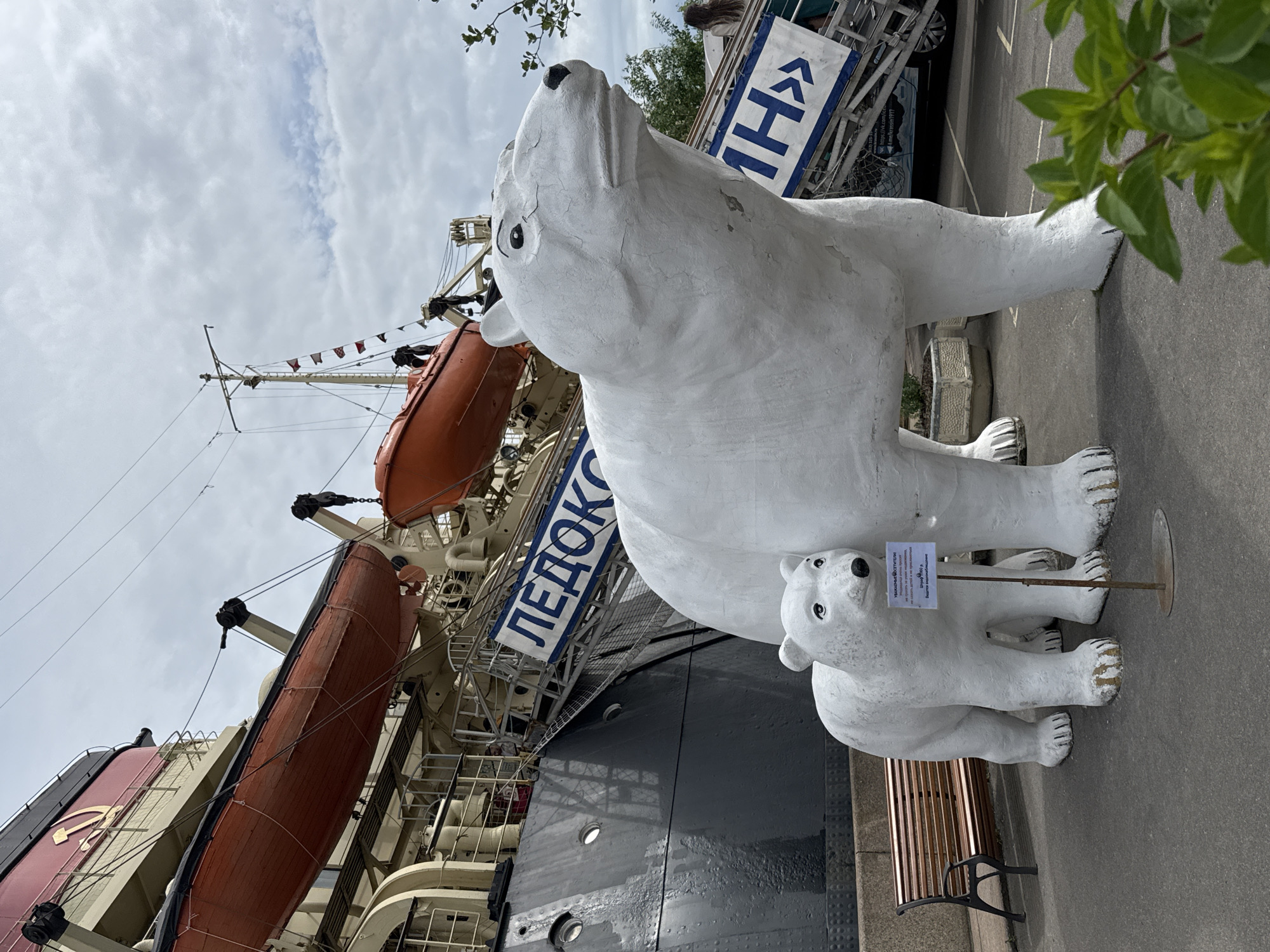 The image depicts the Krassin icebreaker, a museum ship docked in Saint Petersburg, Russia. In the foreground, two white polar bear sculptures stand on asphalt. The larger sculpture is a life-sized representation of an adult polar bear, while a smaller one, about half its size, stands beside it. A small informational sign is placed between the two bear sculptures. In the background, the gray hull of the Krassin icebreaker is visible, along with orange lifeboats suspended from davits. A blue banner with white Cyrillic text reading "ЛЕДОКОЛ" (Icebreaker) is attached to a ramp leading onto the ship. A wooden bench is situated to the left of the polar bear sculptures. The sky is overcast with some clouds visible.