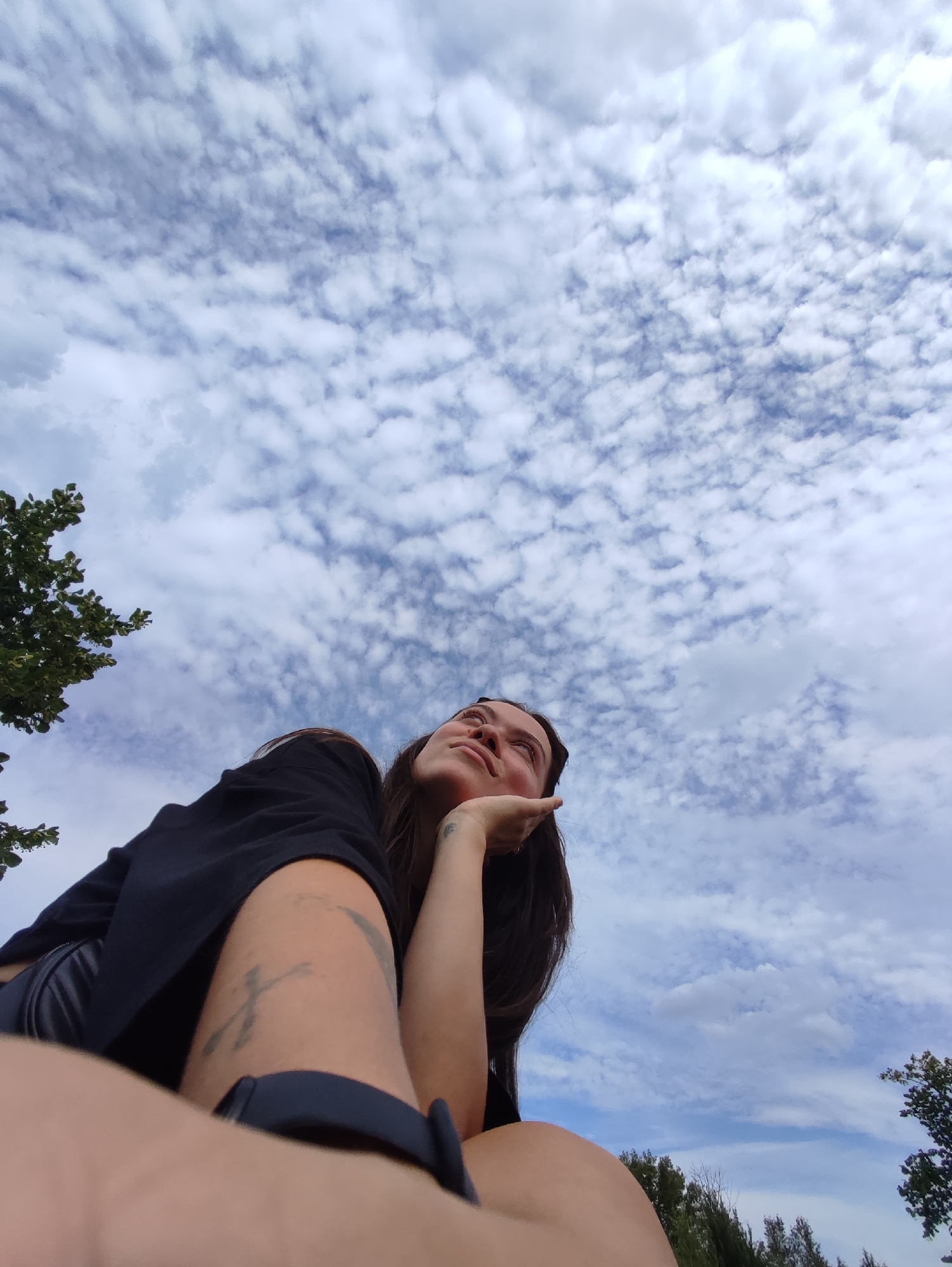 A low-angle shot captures a young woman from the chest up, looking upwards with her hand on her cheek, against a backdrop of a cloudy blue sky. The woman has dark hair and is wearing a black top. A tattoo is visible on her left arm. The scene appears to be outdoors, with trees visible in the lower left and right corners of the frame, suggesting a park or natural setting within the city of Chaikovsky, Russia.
