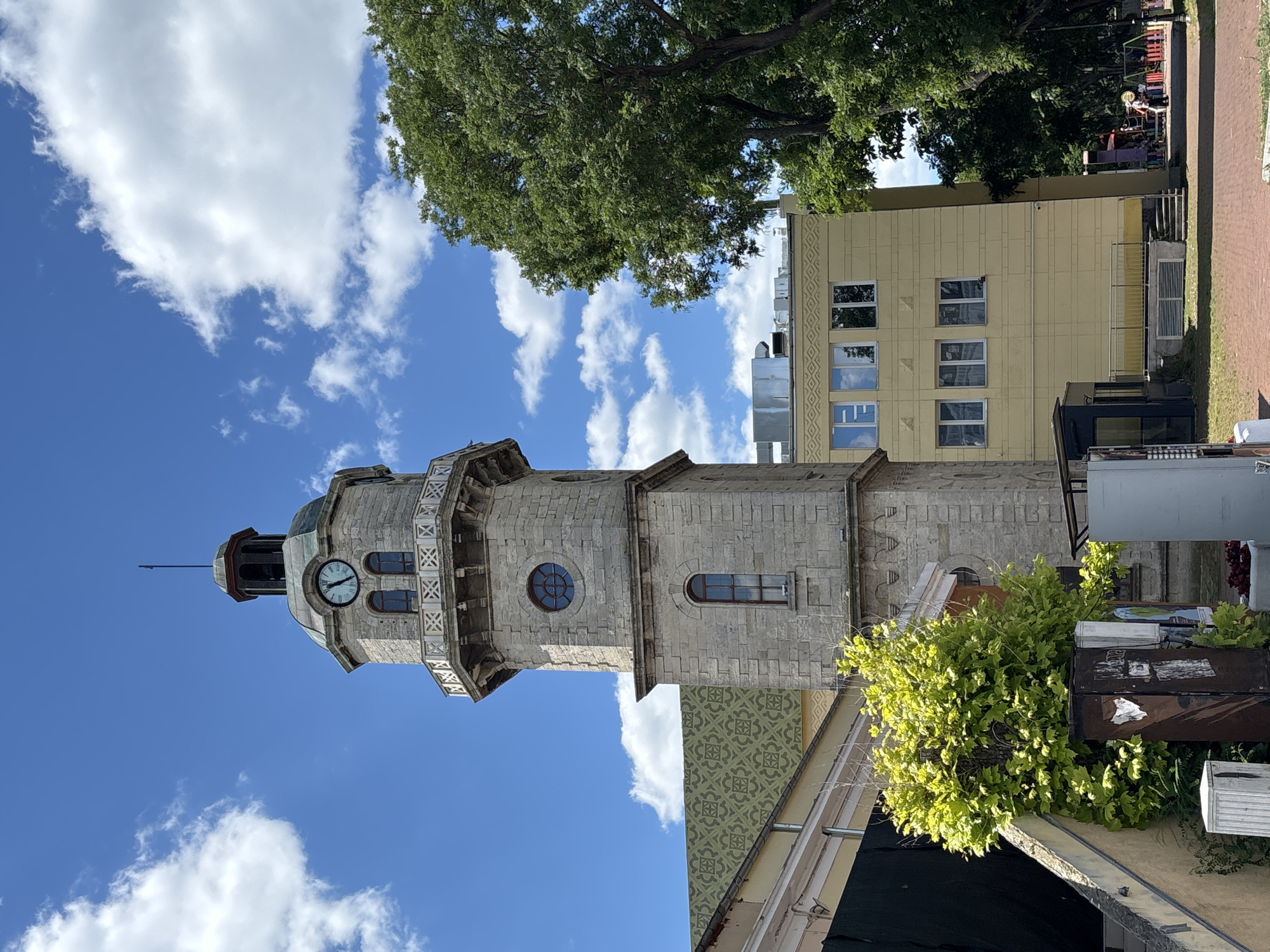 The image captures a view of the Varna Clock Tower, a prominent landmark located in Varna, Bulgaria. The tower is constructed of light-colored stone and features a clock face with Roman numerals. It has an octagonal top section with a small spire. Below the clock, there is a decorative balcony with white railings. The tower is partially obscured by a lush green tree to its right.
Adjacent to the clock tower, a modern, yellow building with multiple windows is visible. The building has a flat roof and an external air conditioning unit. To the left of the clock tower, a building with a green, patterned roof is partially visible. In the foreground, a concrete staircase leads up to a wall covered in dark material, likely a tarp or banner. A vibrant green bush grows beside the staircase, and a weathered metal box is also present.
The sky above is a clear blue, dotted with fluffy white clouds. The ground is paved with reddish bricks, suggesting a public square or walkway. In the background, the dense foliage of trees lines a path, and a small playground with colorful equipment is visible, along with a few indistinct figures of people. The overall scene depicts a sunny day in Varna, highlighting the juxtaposition of historical architecture and modern urban elements.