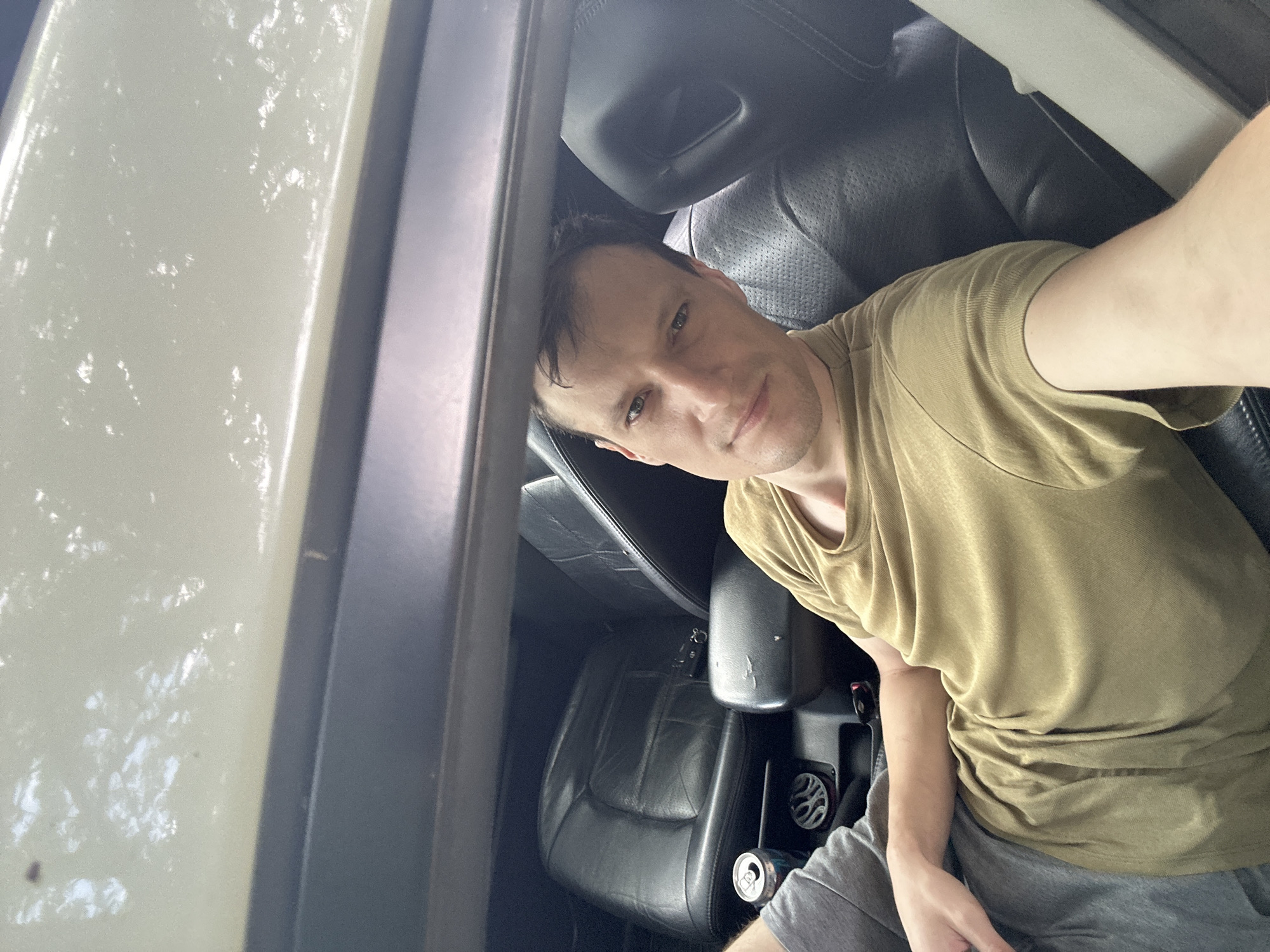 A man is sitting in the passenger seat of a car, his body angled towards the camera. He is wearing a tan t-shirt and grey shorts. The interior of the car appears to be leather, with the driver's seat visible behind him. A can of soda is on the center console. The car appears to be parked outdoors, with sunlight filtering through trees and reflecting on the car's roof, suggesting it is daytime. The location is Makiv, Ukraine.