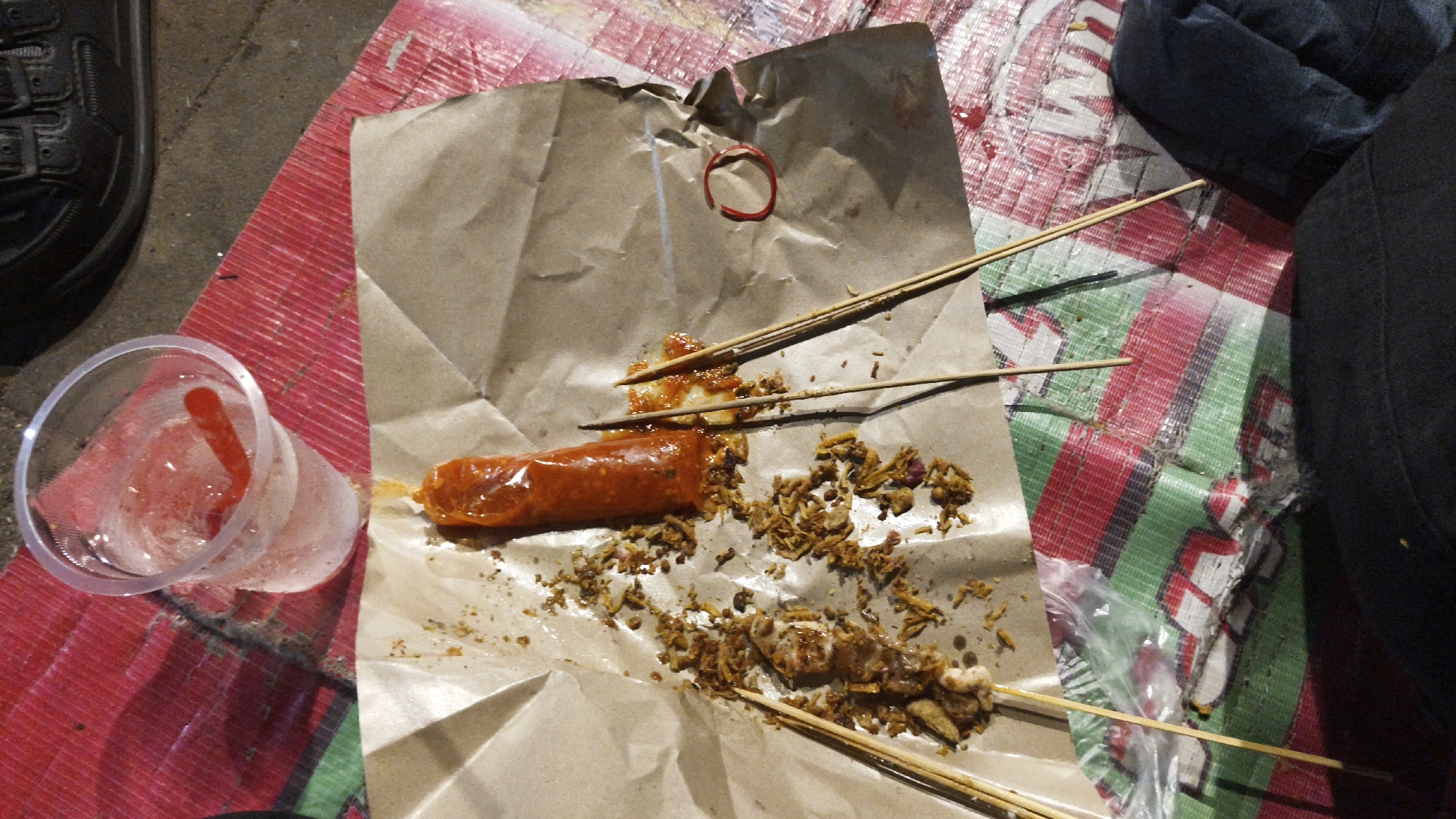 The image depicts a close-up, overhead view of food items scattered on brown paper, likely on a street or casual dining area in South Jakarta, Indonesia. In the foreground, a clear plastic cup filled with ice and a red liquid, possibly a beverage, is visible. To the right, a long, sausage-like food item coated in a bright orange sauce is placed on the crumpled brown paper. Several wooden skewers are interspersed with the food, some holding pieces of meat or other small, dark, crispy food items. The food appears to have been eaten, as evidenced by the scattered remnants. The background shows a red and green striped mat, possibly a tablecloth or ground cover, and a portion of dark footwear, suggesting a person is present but not fully visible. The lighting suggests it is either evening or nighttime.