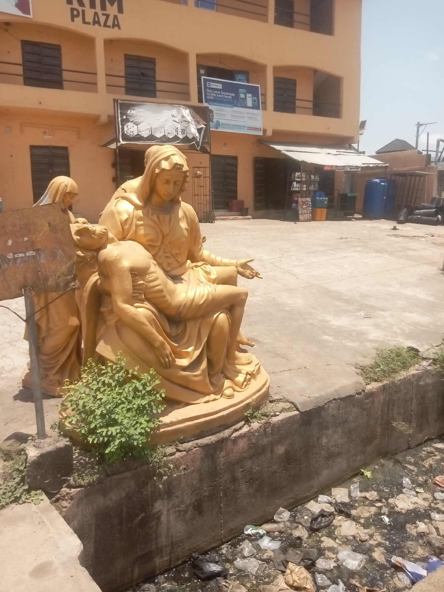 The image captures an outdoor scene in Onitsha, Nigeria, featuring a golden religious sculpture in the foreground, set against a backdrop of a commercial building and an open paved area.
The central subject is a large, golden, Pieta-style sculpture depicting a sorrowful woman, likely Mary, seated and cradling the body of a deceased male figure, presumably Jesus, across her lap. Another female figure stands partially visible to the left, behind the main group. A small, vibrant green bush grows at the base of the sculpture. The sculpture rests on a concrete base, which overlooks a shallow concrete drainage ditch below. This ditch is visibly filled with discarded plastic bottles, bags, and other refuse, alongside murky water, indicating a state of environmental neglect.
In the midground, a wide, open concrete space stretches back towards a commercial building. To the right, a small, informal stall with a corrugated metal roof is set up, displaying various goods for sale. Nearby, several large blue and yellow plastic barrels are stacked, along with bundles of black pipes.
The background is dominated by a two-story commercial building painted in a light peach or brown hue. The building features multiple dark-paned windows and balconies. Prominently displayed near the top of the building are the words "RIM PLAZA." Below this, a large banner on the building's facade reads, "Get your business to the next level with Pointpoint," with smaller text indicating it is "powered by ANCHORAZI PROPERTIES" and includes a phone number. Another smaller banner is partially visible to the left of the main banner.
The scene is brightly lit by what appears to be strong midday sunlight, casting distinct shadows and suggesting clear, sunny weather. The overall impression is one of a religious display juxtaposed with a busy urban commercial setting and some signs of environmental degradation.
