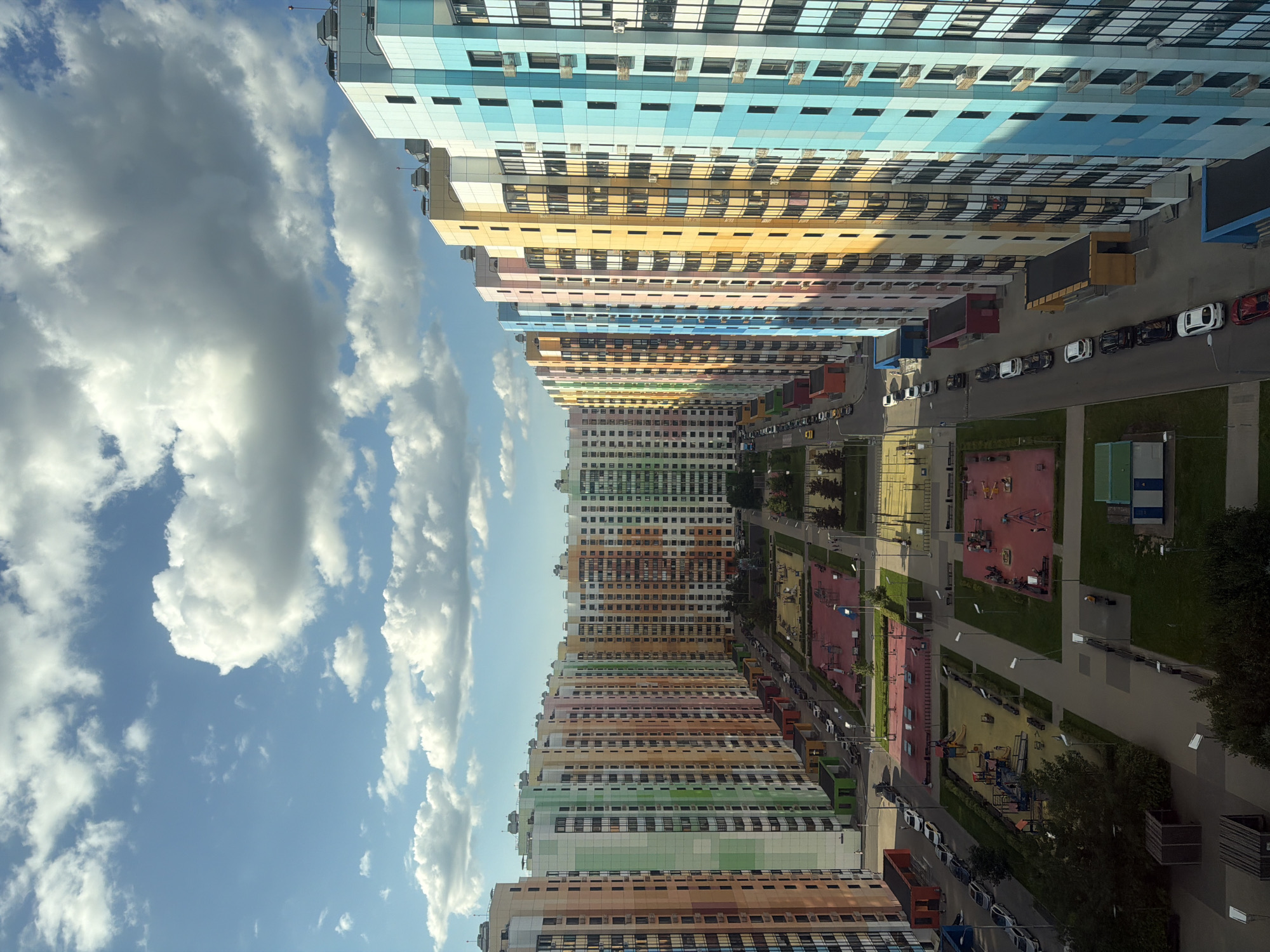 The image displays a high-angle view of a modern residential complex with several tall, multi-colored buildings arranged in parallel rows. The buildings are brightly painted in a spectrum of colors including orange, pink, yellow, blue, and green, with many windows and balconies visible. Between the buildings, there are courtyards with playgrounds featuring red and yellow surfaces, swings, slides, and other play equipment. A paved road runs alongside one row of buildings, with several cars parked along it and on the side. There are also pedestrian walkways, benches, lampposts, and some areas of grass and trees. The sky above is a clear blue with scattered white clouds. The overall impression is of a vibrant and organized urban environment.