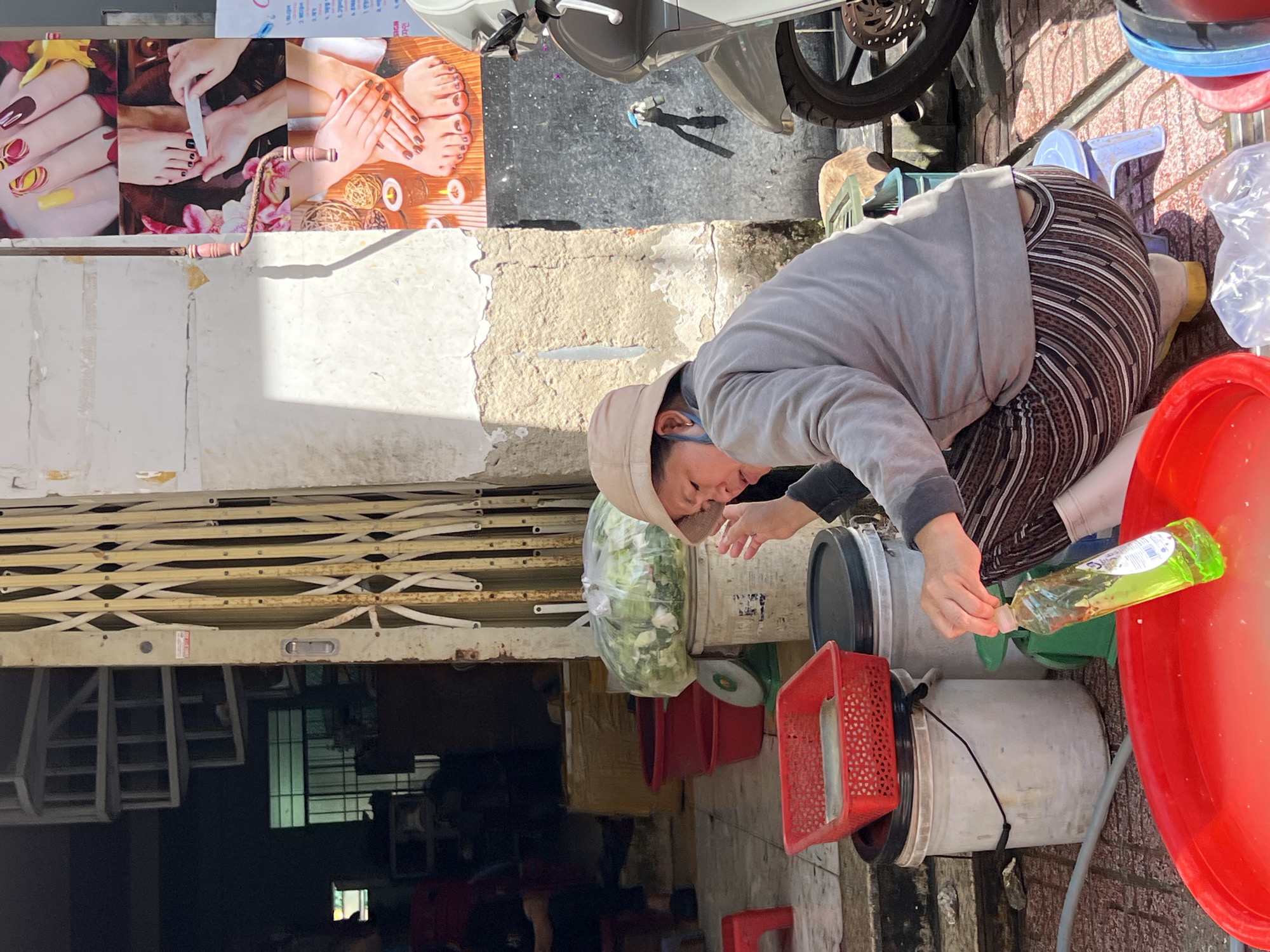 In Nha Trang Ward, Vietnam, a woman wearing a tan hat and a grey hoodie is crouched on the ground next to several large plastic buckets and red baskets. She is holding a clear plastic bottle filled with green liquid, tilting it to pour into a large red basin in the foreground. The woman has on striped pants and yellow footwear. In the background, a metal roll-up gate is partially open, revealing a staircase and a doorway. A large plastic bag filled with green leafy vegetables sits on top of a white bucket with a green scale underneath. Above the woman, a colorful poster advertising nail and foot services is attached to a weathered wall. A portion of a silver scooter is visible to the right of the frame. The ground is paved with reddish-brown tiles, and sunlight casts distinct shadows.