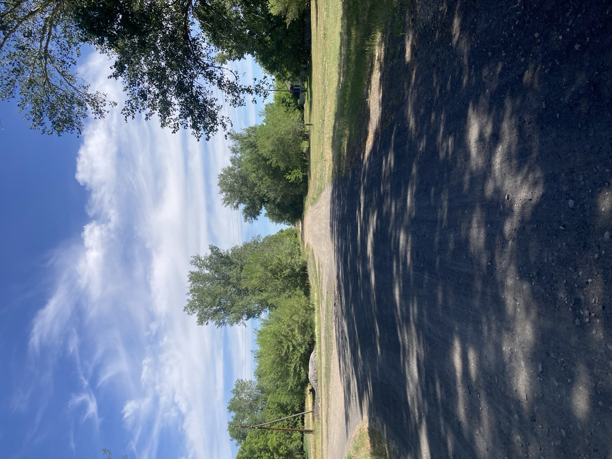 The image depicts a dirt road leading into a rural area. On the left side of the road, there is a power pole and a pile of gravel. The road is lined with trees and green grass. In the distance, there appears to be a house. The sky is blue with scattered white clouds. The sun is casting shadows on the road, indicating it is daytime. The image appears to be taken in the village of Rakity, Russia, given the context provided.