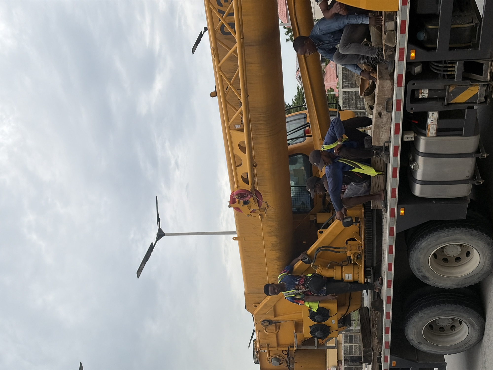 In Alaguntan, Nigeria, a crane truck is parked with its boom extended. Five young men are positioned on the truck bed and on the crane's base. One man, wearing a colorful t-shirt and a high-visibility vest, stands near the crane's cabin. Two men are crouched down on the truck bed, one in a blue denim shirt and the other in a blue shirt with a yellow vest. Another man in a denim shirt is seated on a wooden block on the truck bed. A fifth man, partially visible, is also seated on the truck bed. The sky is overcast, with a tall lamppost visible in the background. The truck appears to be a heavy-duty vehicle, with large tires and a metallic fuel tank. The overall scene suggests a construction or industrial setting.