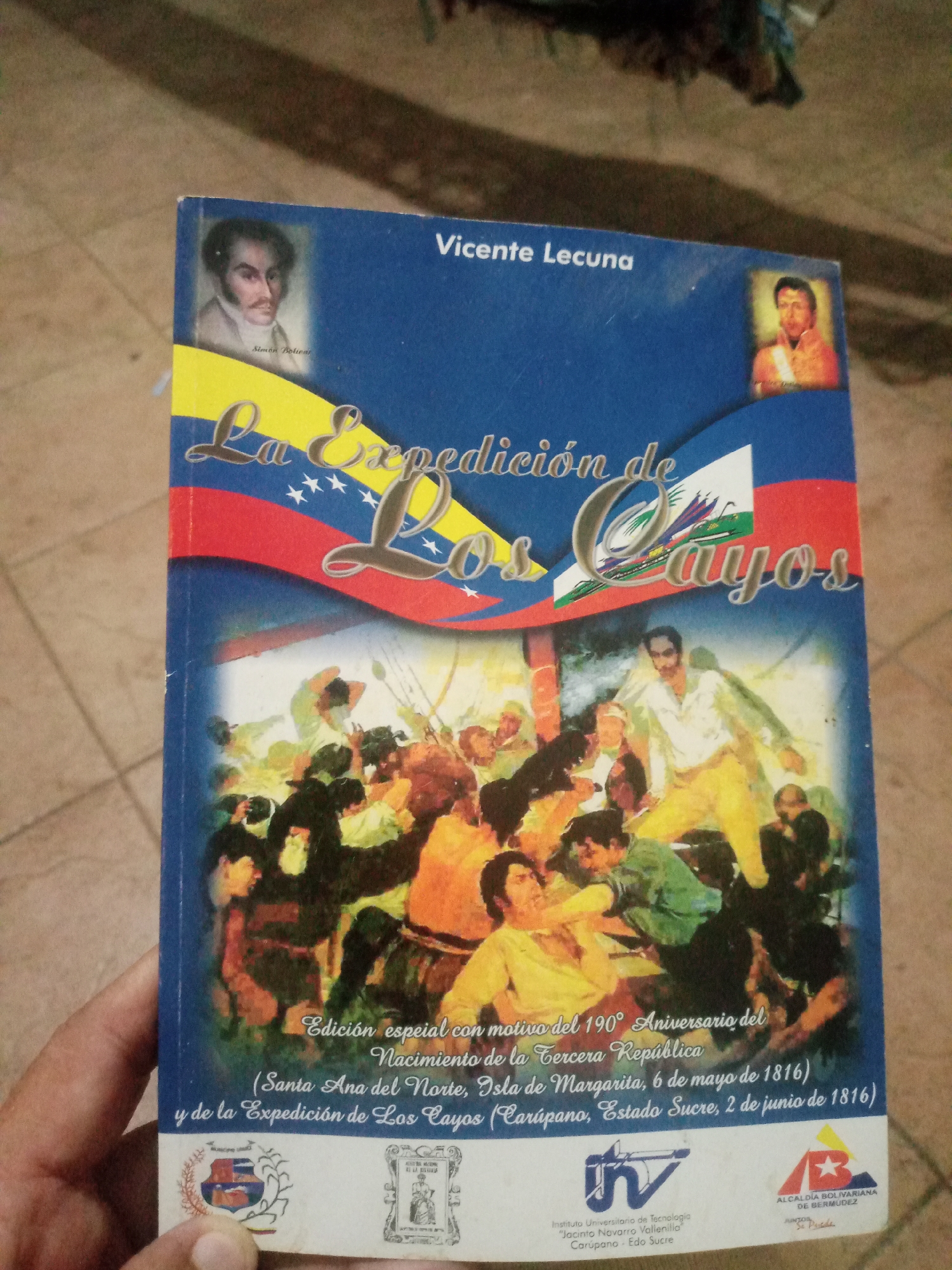 The image shows the cover of a book titled "La Expedición de Los Cayos" by Vicente Lecuna. The cover features portraits of historical figures and a depiction of a historical event. The text on the cover indicates that it is a special edition commemorating the 190th anniversary of the Third Republic, and mentions the city of Santa Ana del Norte, Isla de Margarita, on May 6, 1816, and the expedition to Los Cayos (Carúpano, Estado Sucre) on June 2, 1816. The bottom of the cover displays logos of institutions, including the Instituto Universitario de Tecnología "Jacinto Navarro Vallenilla" in Carúpano, Edo Sucre, and the Alcaldía Bolivariana de Bermúdez. The book is being held in a person's hand, with a tiled floor visible in the background.