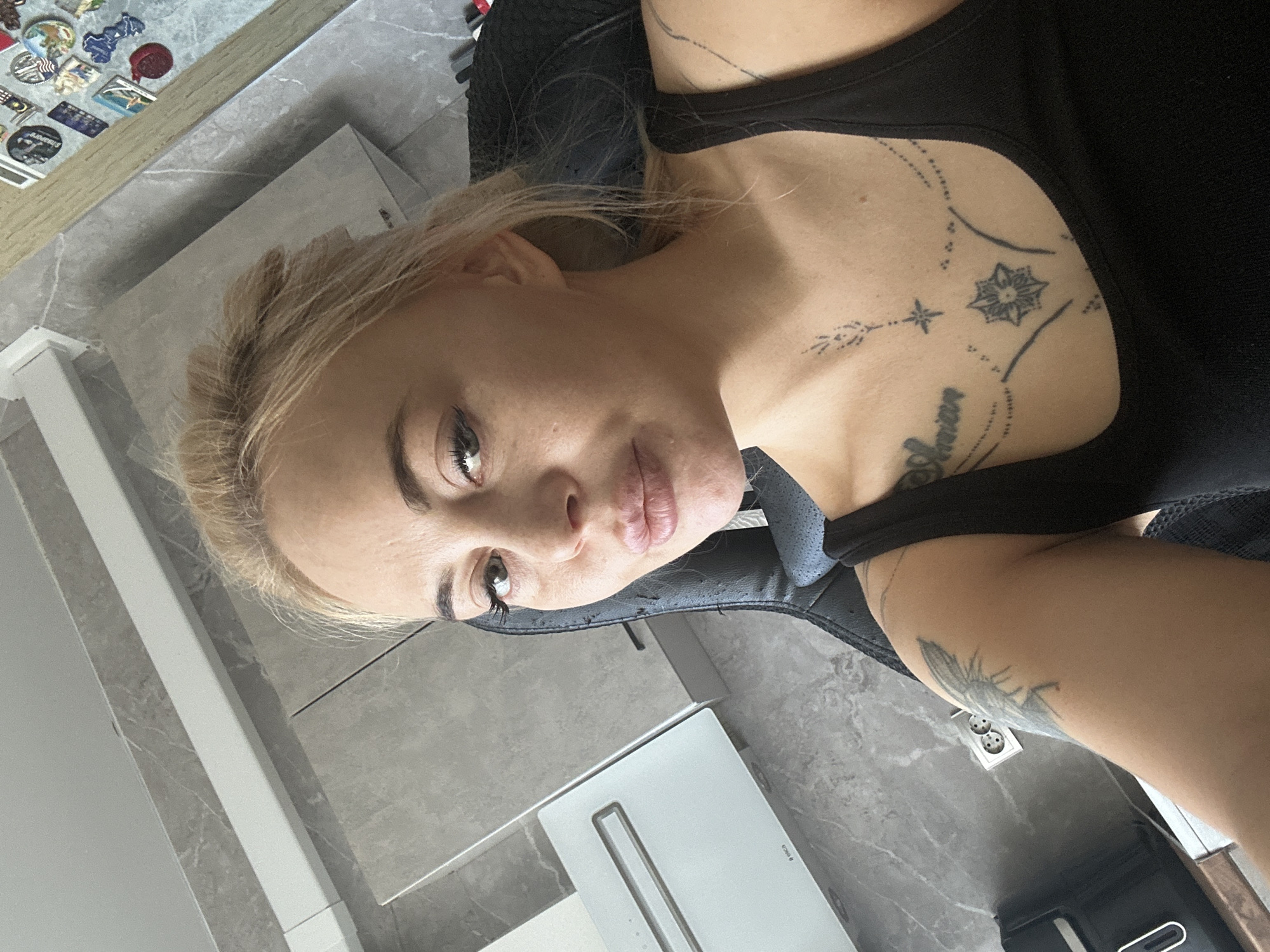 A woman with blonde hair is seated indoors, possibly in Высокий, Russia. She is wearing a black tank top and has tattoos on her chest and arm. The background includes a grey wall, a white cabinet, and a black chair.