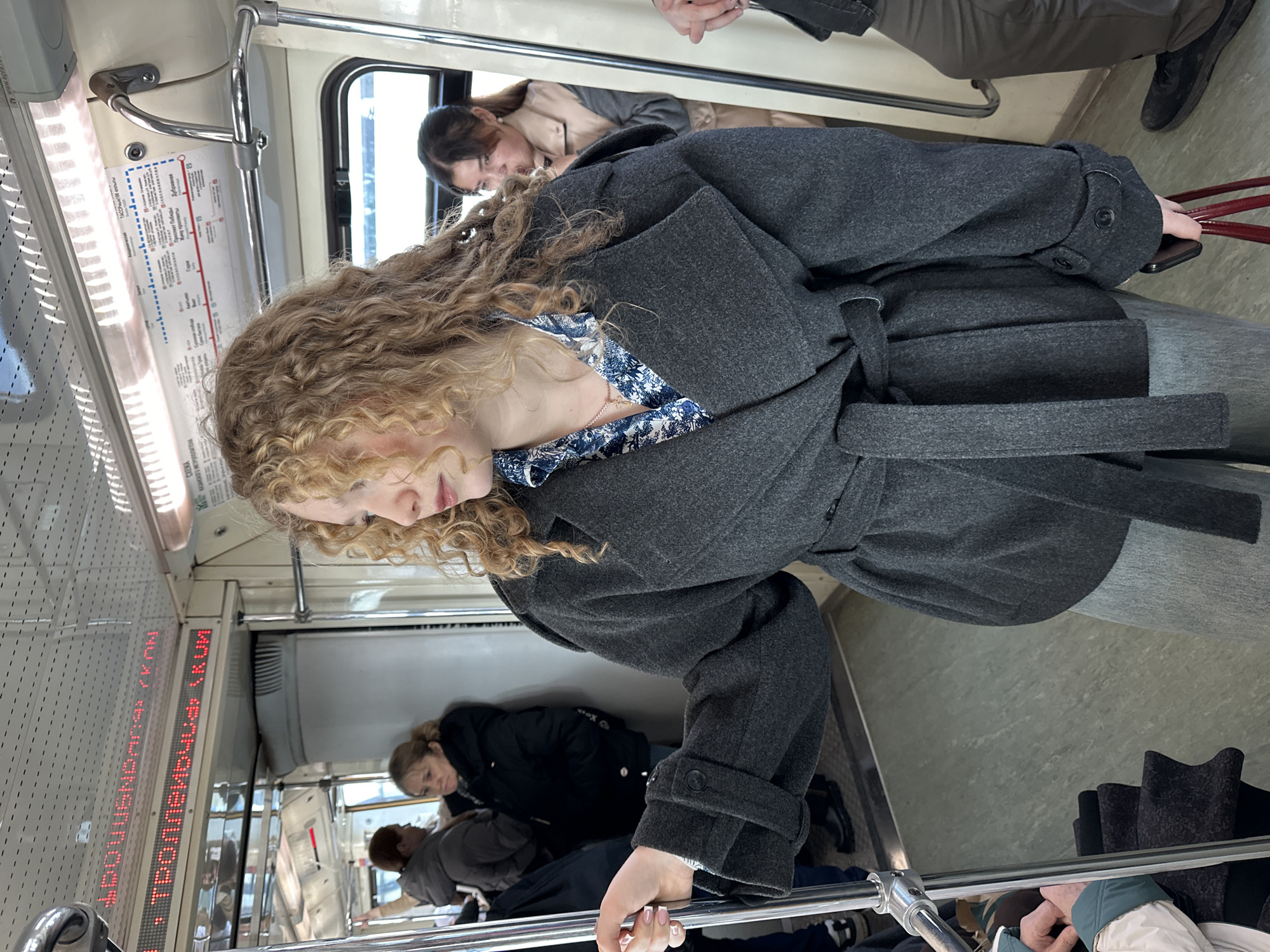 The image displays the interior of a Kazan Metro train car in Kazan, Russia.
A young adult female with light brown curly hair occupies the central foreground, wearing a dark grey belted coat, a blue and white patterned shirt, and light grey trousers. Her left hand is gripping a vertical chrome support pole, while her right hand holds a dark smartphone and a red tote bag. She is looking to her left.
In the left background, two female passengers are visible: one mostly obscured in a dark hooded jacket, and another middle-aged woman in a dark hooded winter coat, facing away from the camera. In the right background, a young woman with dark hair and a light-colored jacket is partially visible through an opening, looking left. The bottom left foreground shows the hands and arm of another passenger on a seat, wearing a white and light blue jacket. The bottom right foreground reveals the legs of a person in dark brown trousers and black shoes.
The train car features cream-colored walls, a pale green floor, and chrome poles. An overhead LED display shows red Cyrillic text, partially legible as "ТРОЛЛЕЙБУСНЫЙ" (TROLLEYBUSNY) and "УЛ. СТОЛБОВАЯ" (STOLBOVAYA ST.). A "МЕТРО КАЗАНЬ" (Kazan Metro) line map, including the station "Дубравная" (Dubravnaya), is mounted above the windows on the right. Overhead lighting is visible, casting reflections. Passengers are standing, indicating movement or waiting within the metro system.