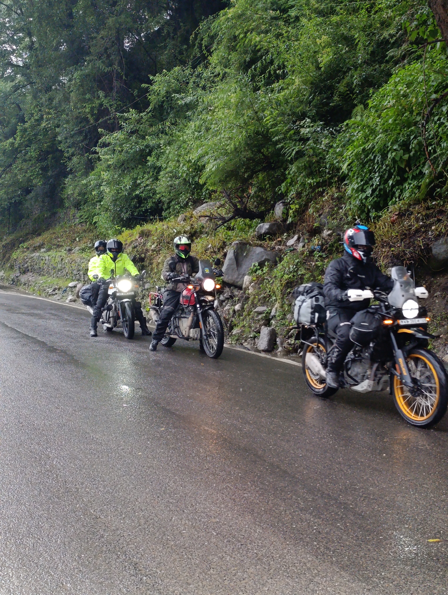 A group of four motorcyclists, wearing helmets and riding gear, are traveling on a wet asphalt road. The road is bordered by a steep, tree-covered embankment on the right and a rocky, vegetated slope on the left. The motorcyclists are spaced out, with the lead rider on a dark motorcycle with yellow accents, followed by two riders on a lighter-colored motorcycle, and another rider on a red motorcycle with yellow wheel rims. The sky appears overcast, and the road surface reflects the ambient light, indicating recent rain or damp conditions. The scene is consistent with a motorcycle tour in a mountainous region, likely in Vashisht, India, given the lush vegetation and road conditions.