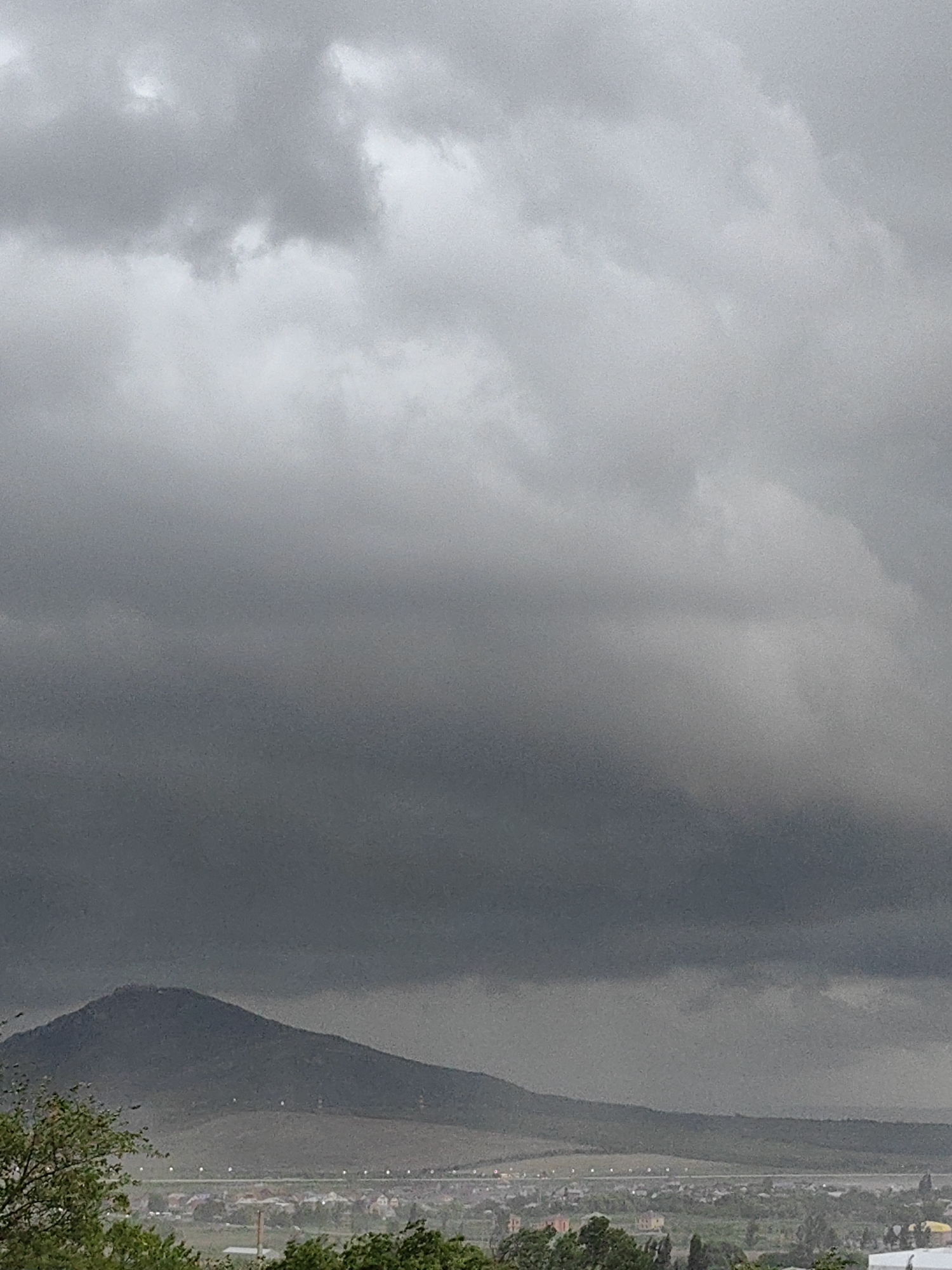 A wide shot captures a panoramic view of a vast, overcast sky dominating the upper half of the frame. The clouds are heavy and dark, suggesting an impending storm. Below the cloudy expanse, a prominent, dark mountain range occupies the middle ground, its peak reaching towards the ominous sky. In the foreground, a lush green landscape of trees and vegetation creates a stark contrast with the brooding atmosphere above. Beyond the trees, a town or village is visible, with its buildings and structures scattered across the land, appearing small and distant. The overall scene evokes a sense of impending weather change and a serene, yet potentially dramatic, natural environment. The location is identified as Unknown, Unknown.