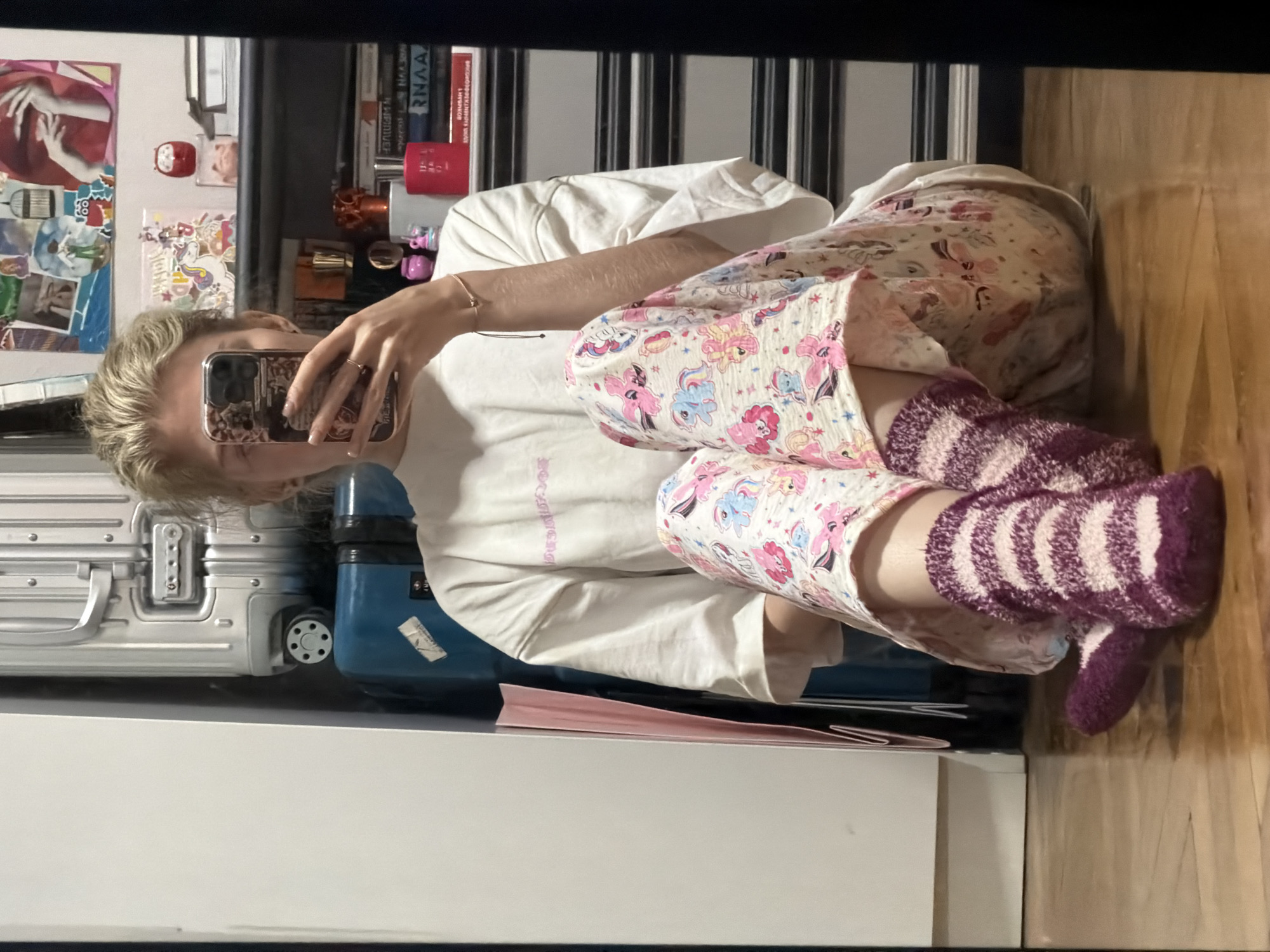A young blonde woman is sitting on a wooden floor, reflected in a mirror. She is wearing a white t-shirt and pajama pants patterned with cartoon unicorns. Her feet are adorned with fuzzy, striped socks in shades of purple and pink. In the background, luggage, shelves filled with books and knick-knacks, and a glimpse of a white wall are visible. The reflection suggests the image was taken in an indoor setting, possibly a bedroom or living area. The presence of luggage might indicate a home preparing for or returning from travel.