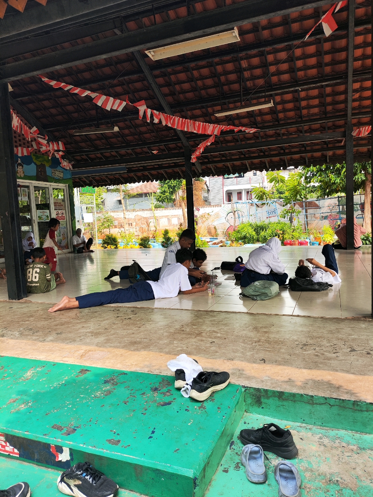 In East Jakarta, Indonesia, within an open-air pavilion adorned with red and white Indonesian flags, a group of students are engaged in various activities on the tiled floor. One student, wearing a white shirt and blue pants, lies prone with their legs extended, appearing to be looking at something on the ground. Nearby, another student in similar attire is seated with their back to the camera, looking down at a device. Further to the left, a student in a green shirt with the number "86" and "NEW YORK" printed on it sits on the floor. To the right, a student in a white hijab and blue pants is also seated and looking at a device. Behind them, other individuals are visible, some seated and others standing. The pavilion's roof is constructed with red tiles supported by dark metal beams, and fluorescent lighting fixtures are installed overhead. The background reveals a garden area with green foliage, colorful flower pots, and a wall decorated with murals depicting cartoon characters and text. In the foreground, a raised green platform displays several pairs of shoes, including black sneakers and light blue sandals. The overall scene suggests a casual break or study session among students in an outdoor school or community setting.
