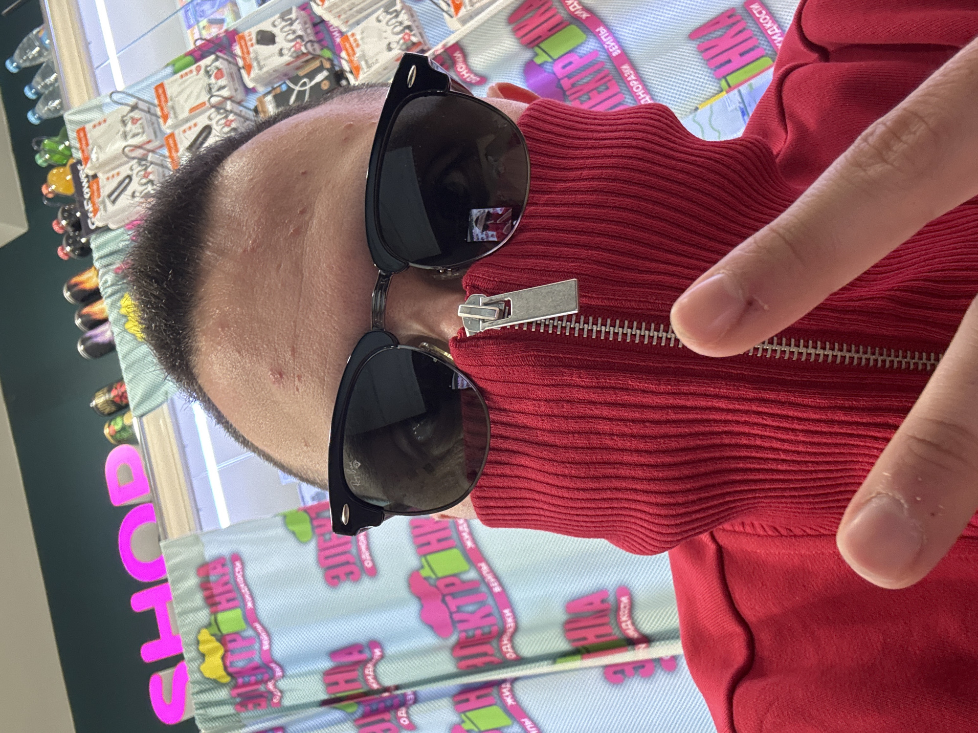 A close-up shot depicts a person wearing sunglasses and a red ribbed top with a zipper. The person's finger is visible near the zipper. The background shows a display with text in Russian, suggesting the location may be in Russia. "SHOP" in large pink letters is also visible. Based on the Russian text and pink "SHOP" letters, it appears this photograph was taken at the Elektra shop in the city of Saint Petersburg, Russia.