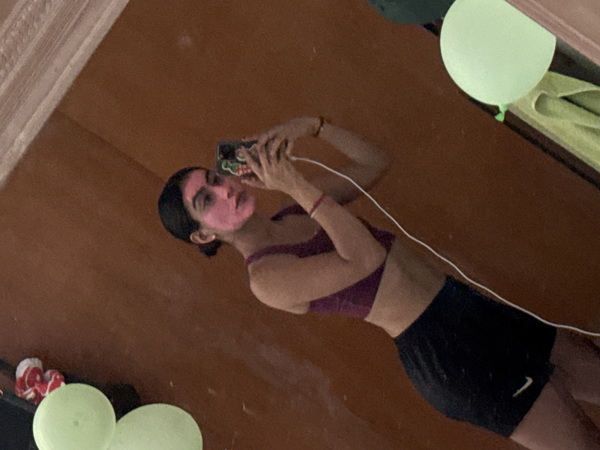 A mirrored image shows a woman in a purple sports bra and black Nike shorts taking a selfie with her phone. She has a pink substance on her face, possibly a mask. A white charging cable is plugged into the phone. There are green balloons scattered in the room. The location details (city of Puebla, Mexico) are not discernible from the visual content of the image.