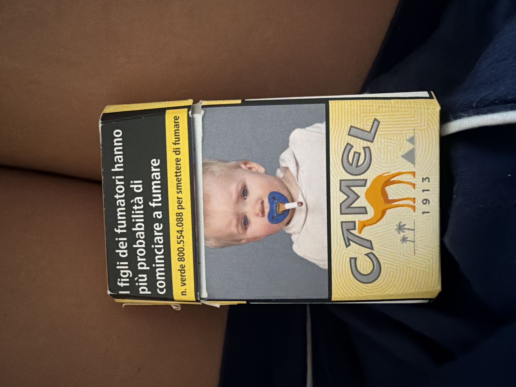 A cigarette pack is held by an unseen person. The pack is predominantly yellow and features a picture of a baby with a pacifier in its mouth, with a lit cigarette sticking out of the pacifier. The text on the pack is in Italian and includes a warning about the increased probability of children of smokers starting to smoke and a phone number for a smoking cessation service. The background is out of focus, showing parts of a person's body and a dark blue fabric. There is no discernible location in Milan or any other city.