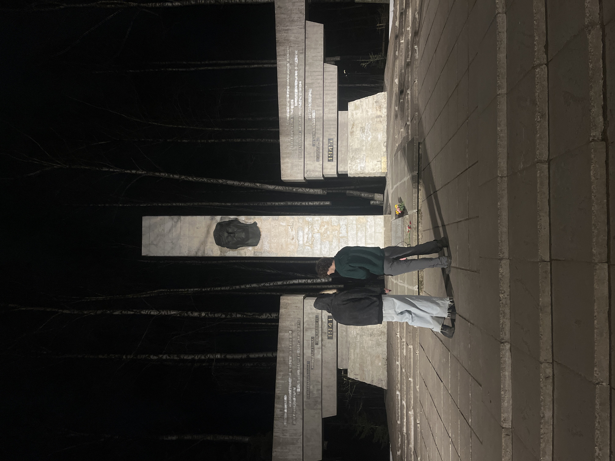 Two people are standing in front of a memorial in Saint Petersburg, Russia at night. The memorial consists of tall, gray concrete slabs with writing on them, and a central panel with a dark sculpture of a person's face. The trees can be seen in the background on the left side. The walkway leading up to the memorial is made of gray concrete blocks. The first person, on the bottom, is wearing a dark jacket and light jeans. The second person, on the top, is wearing a green jacket and gray pants. There are flowers near the edge of the memorial.