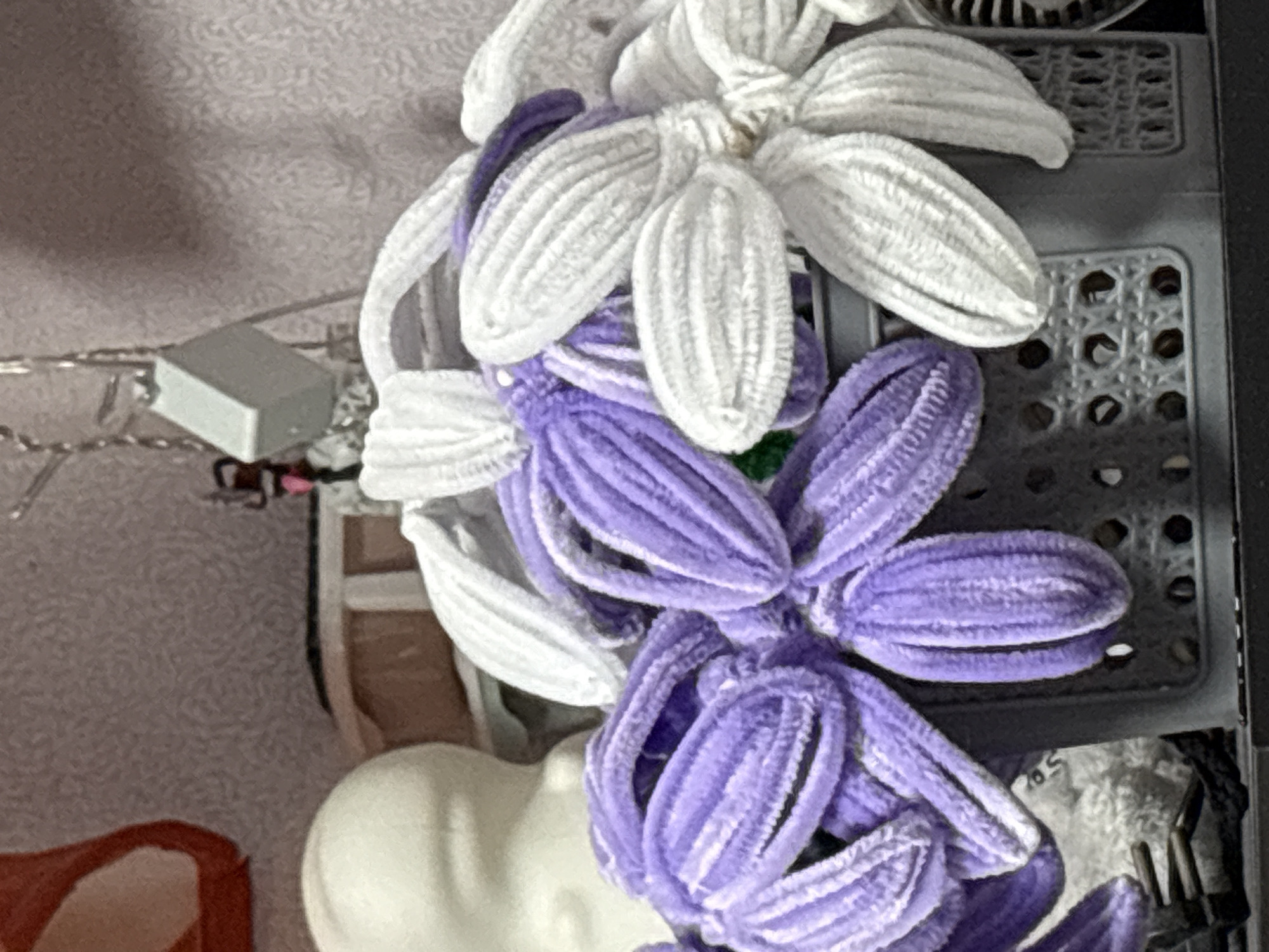 The image features two handcrafted flowers made of what appears to be pipe cleaners. One flower is white, and the other is a lighter shade of purple. The flowers are placed on a gray plastic tray or rack with a grid pattern. To the left of the flowers, there is a small, gray, rectangular object with wires. The background shows a blurred interior space, potentially a room, with hints of furniture and other items. The location in Прикамский, Russia, is not verifiable from the provided image.