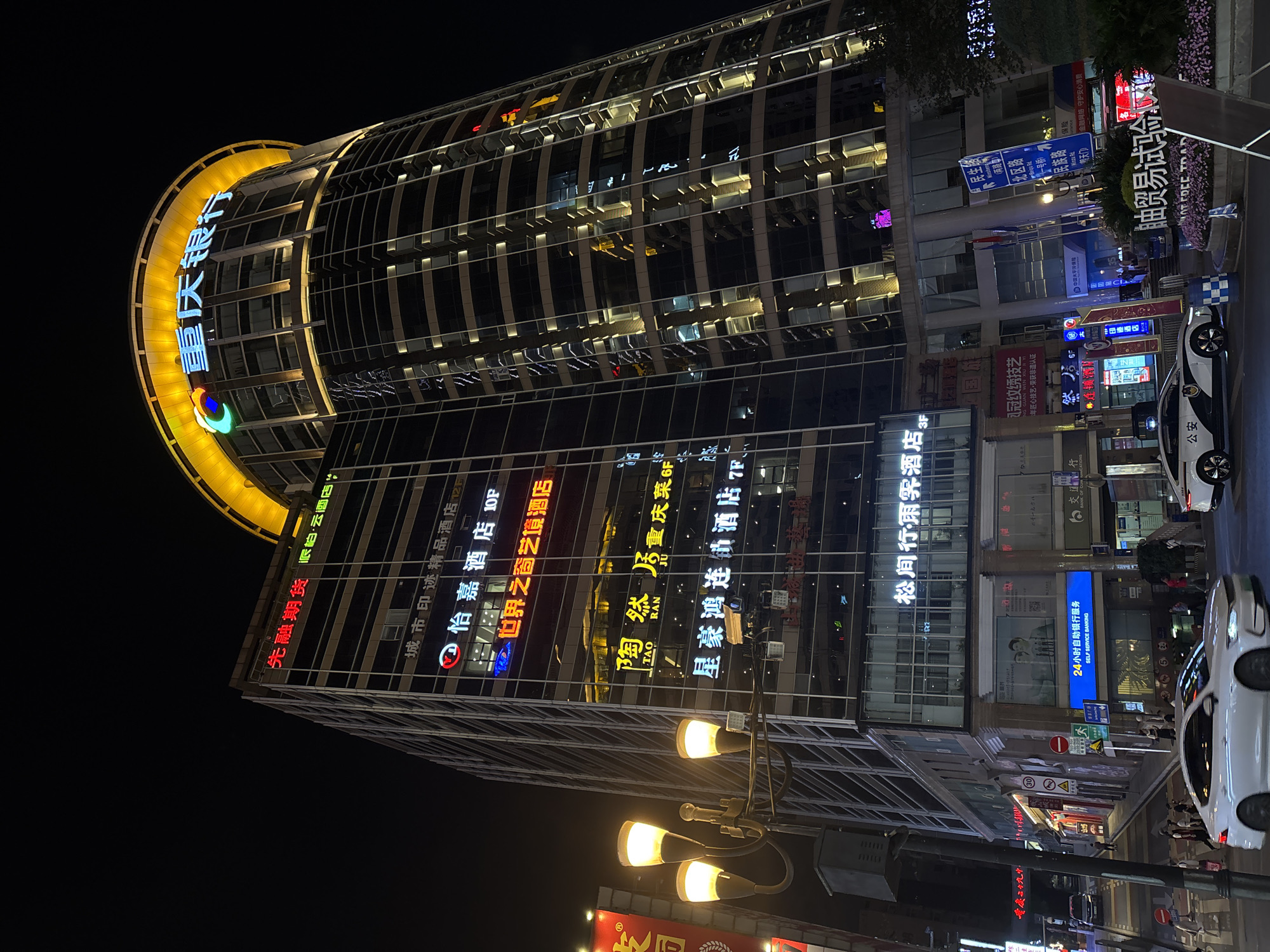 The image shows a nighttime urban scene in Yuzhong District, Chongqing, China. A tall, modern building dominates the view, featuring a cylindrical structure with a brightly lit yellow ring at its top. The characters "重庆银行" (Chongqing Bank) are prominently displayed in blue along the ring. The building's glass facade reflects numerous illuminated signs and advertisements in Chinese, including restaurant names such as "陶然居重庆菜" (Tao Ran Ju Chongqing Cuisine), hotel names such as "怡嘉酒店" (Yi Jia Hotel) and "星豪鸿连劝酒店" (Xinghao Honglianquan Hotel), and financial advertisements like "先融期货" (Xianrong Futures). A street-level view reveals multiple signs, including "刘冠纹绣技艺" (Liu Guan Wenxiu Art), a sign identifying 24-hour self-service banking, and road signs with street names like "民生路" (Minsheng Road) and "民族路" (Minzu Road) written in Chinese. Street lamps with multiple light bulbs illuminate the area. Cars are parked along the street, including a police car.