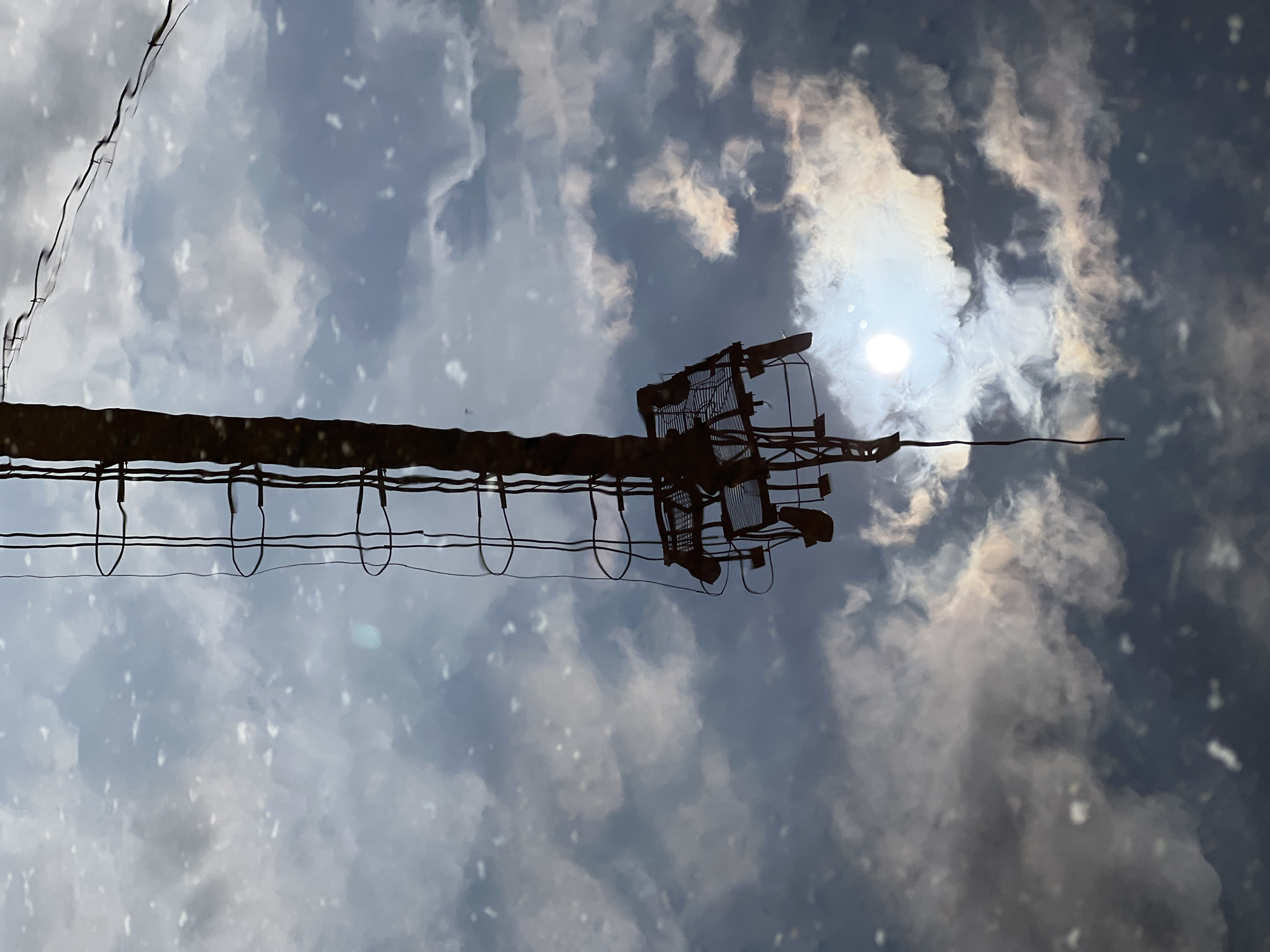 A reflection in a puddle shows a dark, upright structure resembling a ladder against a sky with clouds and the sun. The structure appears to be metallic with rungs and some sort of cage-like attachment near the bottom. The reflection is distorted by ripples in the water. The scene is dark and moody, with the bright sun reflecting in the water adding a point of contrast. The setting appears to be an outdoor urban environment in Veteran-2, Russia, possibly near industrial or utility infrastructure. No people are visible.