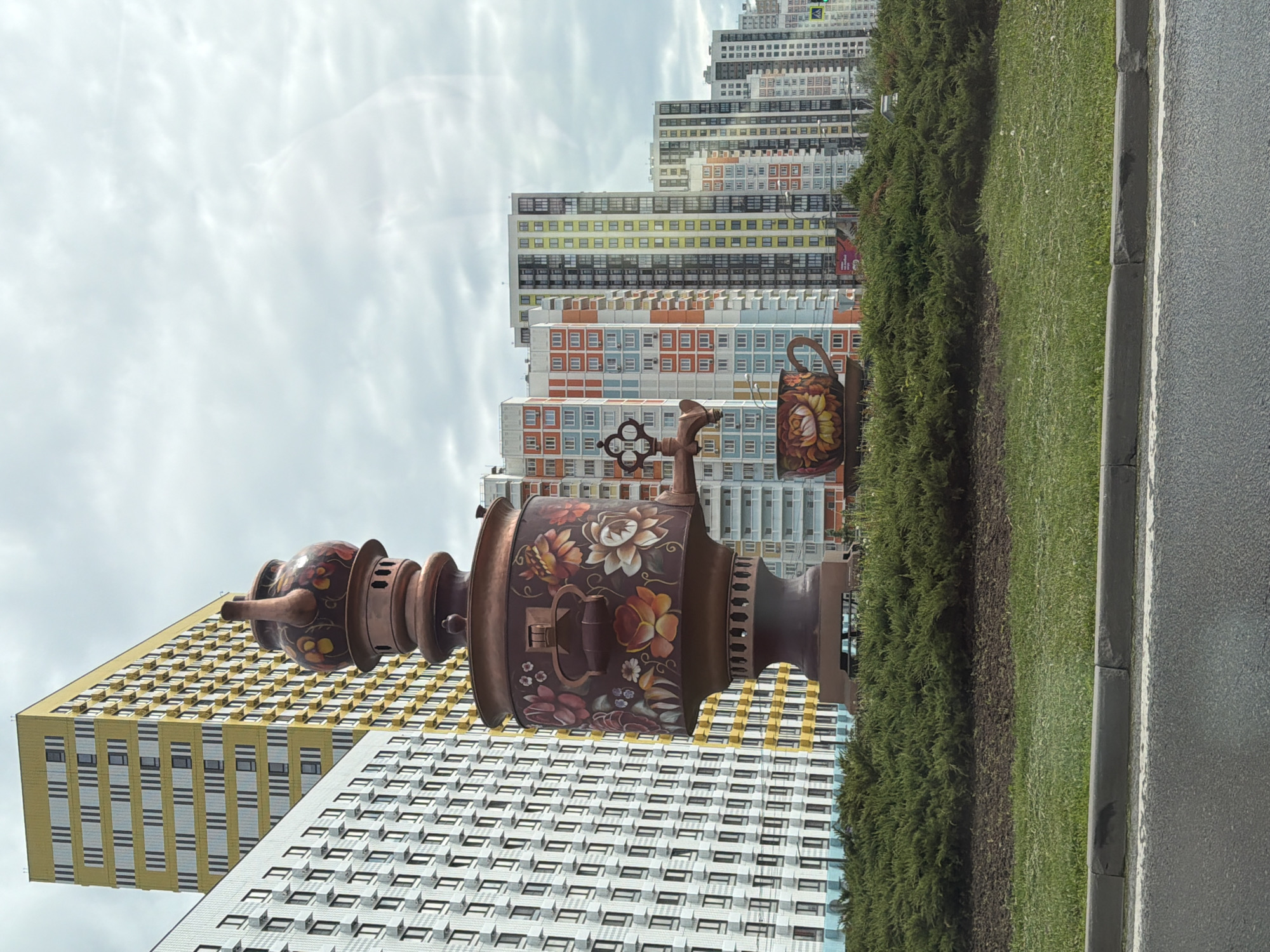 The image shows a large, decorative samovar and teapot sculpture in front of several high-rise apartment buildings. The samovar is painted with floral designs. A green hedge runs along the right side of the frame next to an asphalt road. The sky is overcast.