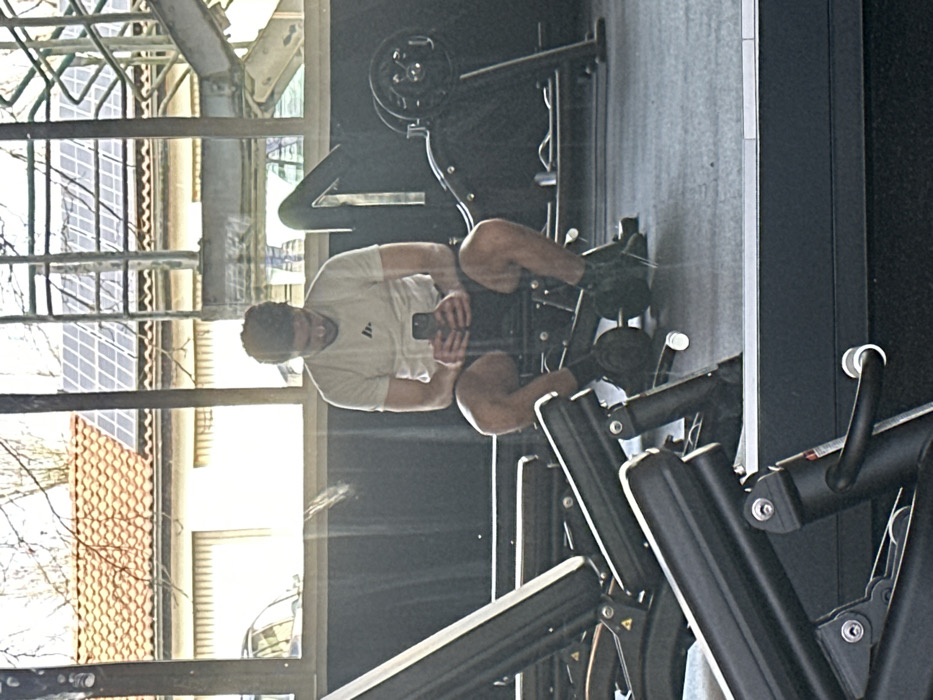 The image depicts the interior of a fitness facility in Jestetten, Germany, with a large window looking out onto an exterior scene. A male subject, appearing to be in his twenties or thirties, is seated on a black padded gym bench, central to the frame. He wears a light-colored t-shirt with a dark logo on the left chest, dark shorts, dark socks, and dark athletic shoes. He is holding a black smartphone with both hands and is looking intently at its screen.
To the subject's right, a black gym machine with a loaded circular weight plate is visible. In the left foreground, components of another black gym machine, including padded sections, are partially visible. The floor is dark grey gym matting.
Through the large window, an exterior environment is visible. A light-colored building with an orange-tiled roof, featuring a prominent array of solar panels, stands in the background. Bare tree branches are discernible against a bright, overcast sky. A metallic structure, possibly a railing or staircase, is also visible outside the window, alongside a partial view of a vehicle. The window surface shows reflections of the gym's interior.