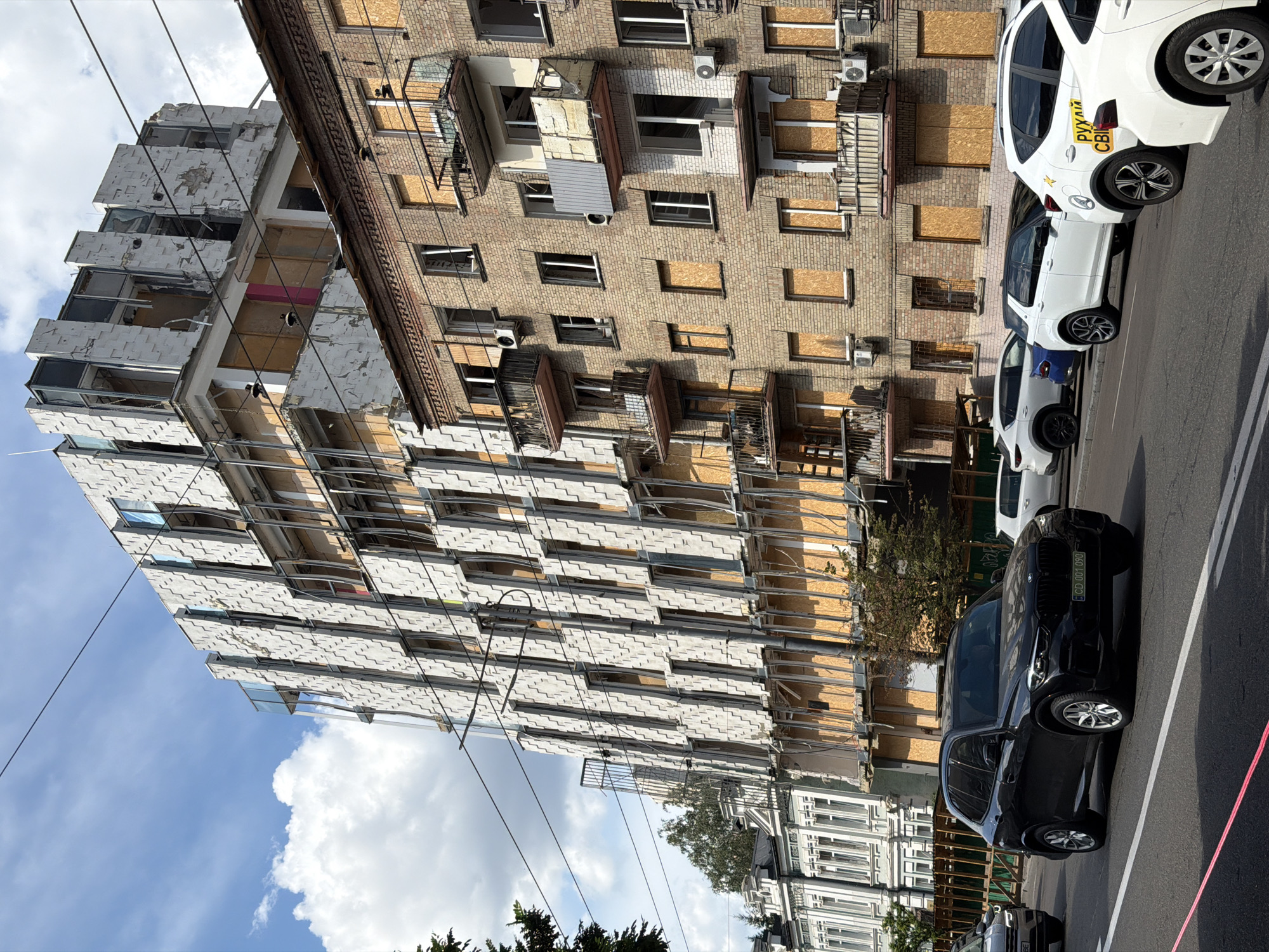 The image shows a street scene in Kyiv, Ukraine, with two multi-story buildings. The building on the left is modern with a distinctive facade of angled white panels, but it appears to be damaged, with some panels missing or askew, and windows boarded up. The building on the right is older, constructed of brick, and also shows signs of damage with many windows boarded up with plywood. Balconies on the brick building are visible, some appearing weathered and damaged. Several cars are parked or driving on the street in the foreground, including a black BMW SUV with a license plate visible. Overhead power lines are present. The sky is partly cloudy with blue patches. The overall impression is one of a city affected by conflict or damage, with ongoing repairs or damage assessment evident.