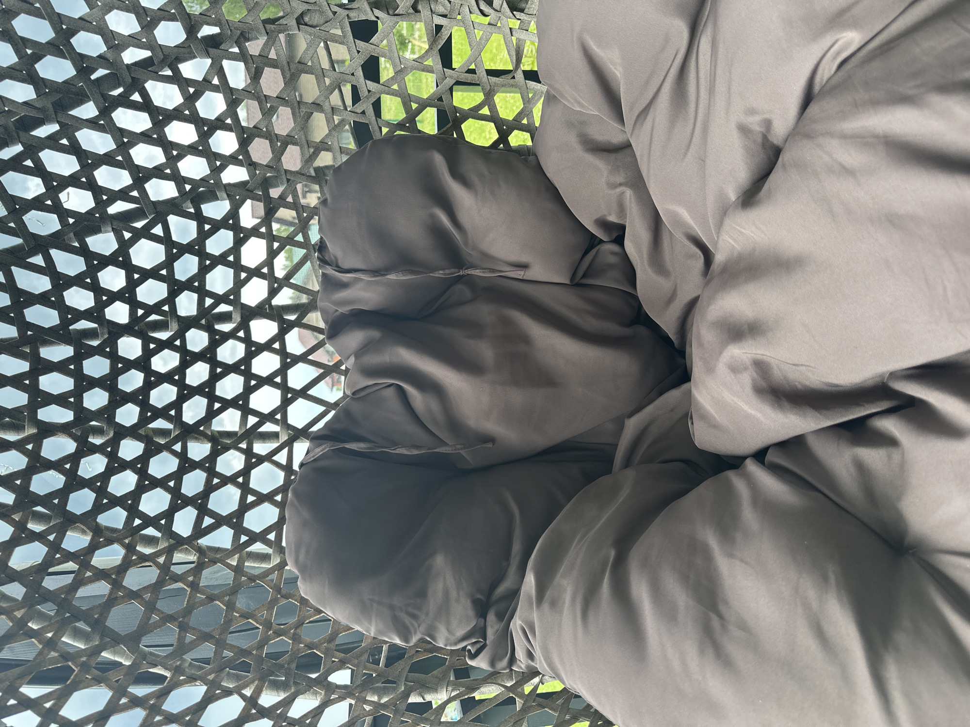 The image shows a close-up view of a dark gray, plush cushion inside a woven hanging chair. The chair's intricate, diamond-shaped weaving is visible in the background, allowing a glimpse of the outdoors. Through the weave, one can see a cloudy sky and hints of houses with tiled roofs, along with green foliage, suggesting a residential area. The cushion is heavily wrinkled and appears to be partially enveloping the viewer, creating a sense of comfort and enclosure. There are no people visible in the image. The setting appears to be outdoors, possibly on a porch or patio, in the city of Chernyshnya, Russia.