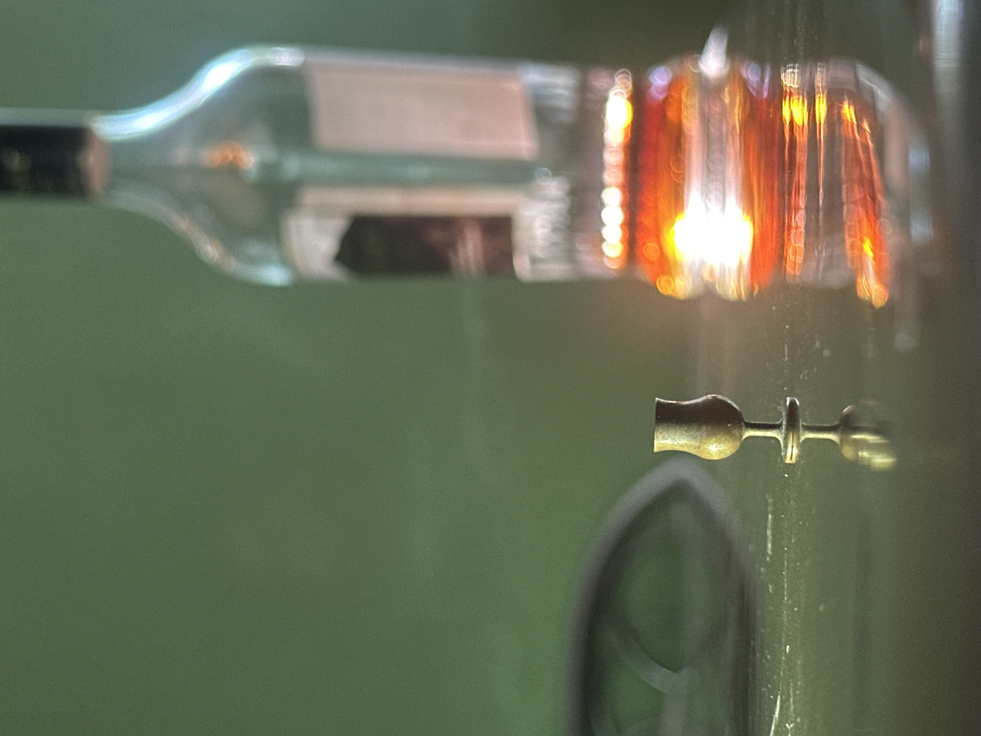 The image depicts a still life composition on a dark, reflective surface. In the foreground, a small, metallic goblet with a delicate stem stands upright, casting a clear reflection. To the right and further back, a clear glass bottle is visible, containing an amber liquid that is illuminated by a bright light source. This light creates a bokeh effect of circular highlights on the bottle's base and on the reflective surface. The background is a solid, dark green color, creating a stark contrast with the subjects. The scene is devoid of any human presence or activity. The location in Lobacheve, Ukraine is not discernible from the visual information provided in the image.