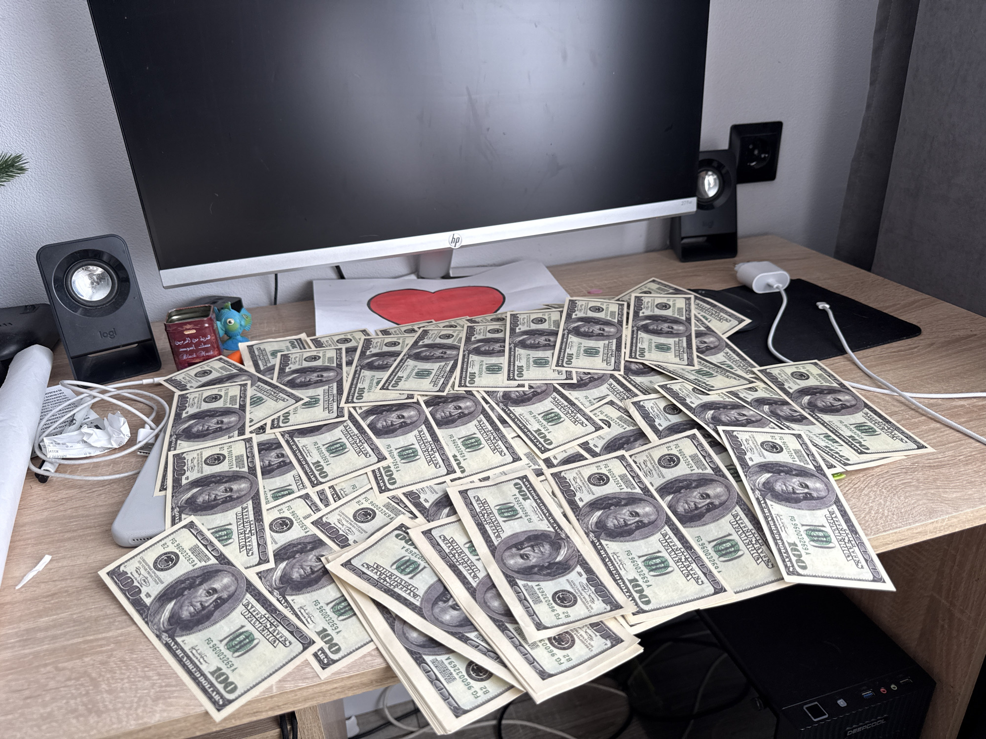 A photograph depicts a light brown wooden desk in an indoor setting. On the desk is a pile of what appear to be numerous United States one hundred dollar bills, scattered across the surface. Centered behind the bills is a computer monitor with the HP logo visible near the bottom. A black Logitech speaker is positioned to the left of the monitor, with another similar speaker on the right. Behind the speakers, against the wall, are electrical outlets.
Additional objects on the desk include a small decorated box, a white phone or electronic device to the left of the money, a white charger, and a black mousepad with connected cable. A sheet of paper with a red heart drawn on it is visible underneath the monitor.
The overall impression is that the desk is cluttered. To the side of the desk, partially obscured, is a black computer tower marked with "DEEPCOOL". The location or time frame cannot be determined from the image alone.