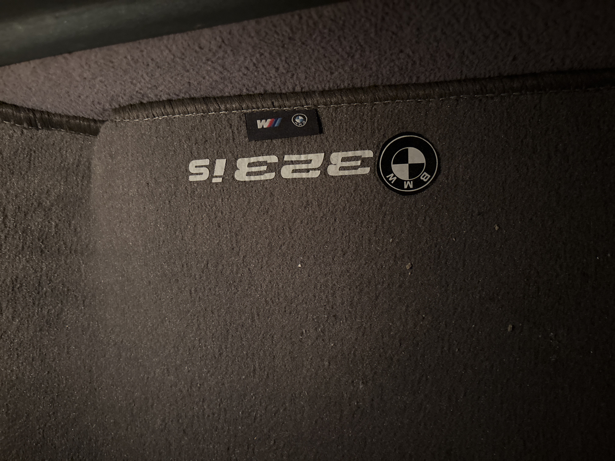 The image depicts a dark grey car floor mat. The mat features white text that reads "323is" in reverse, along with a BMW logo and an "M" logo. The location in the city of Unknown, Unknown is undeterminable.