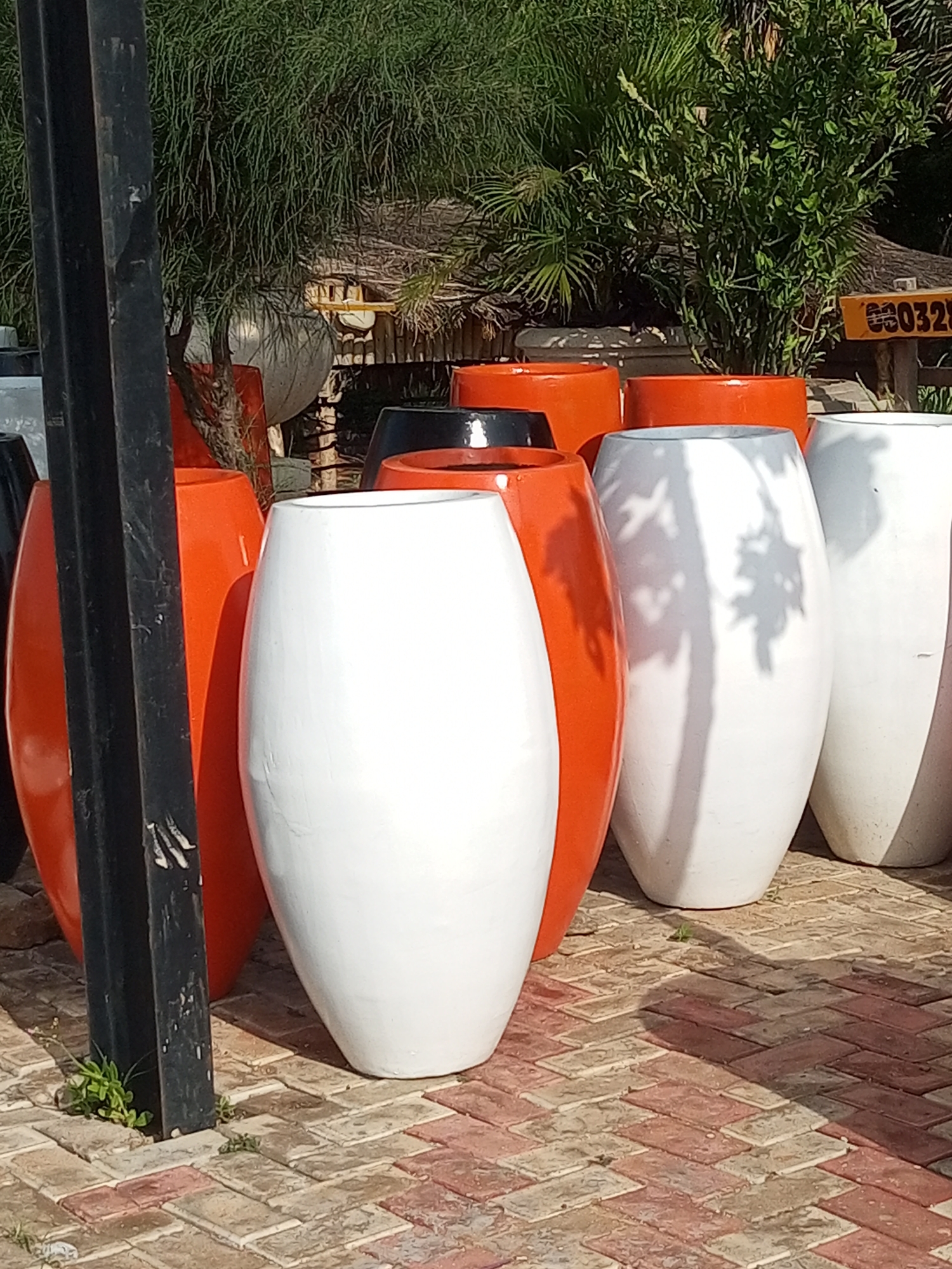 This image shows a collection of large, modern, oval-shaped planters arranged on a patterned brick walkway. The planters are primarily white and orange, with one black planter partially visible. The background features lush green foliage and a thatched roof structure, suggesting an outdoor setting possibly related to landscaping or a garden center. A dark, vertical pole is visible on the left side of the frame, partially obscuring some of the planters. The lighting indicates a sunny day, casting shadows from the foliage onto the planters. There are no people or animals visible in the image. The location is identified as the city of Gombe, Nigeria.