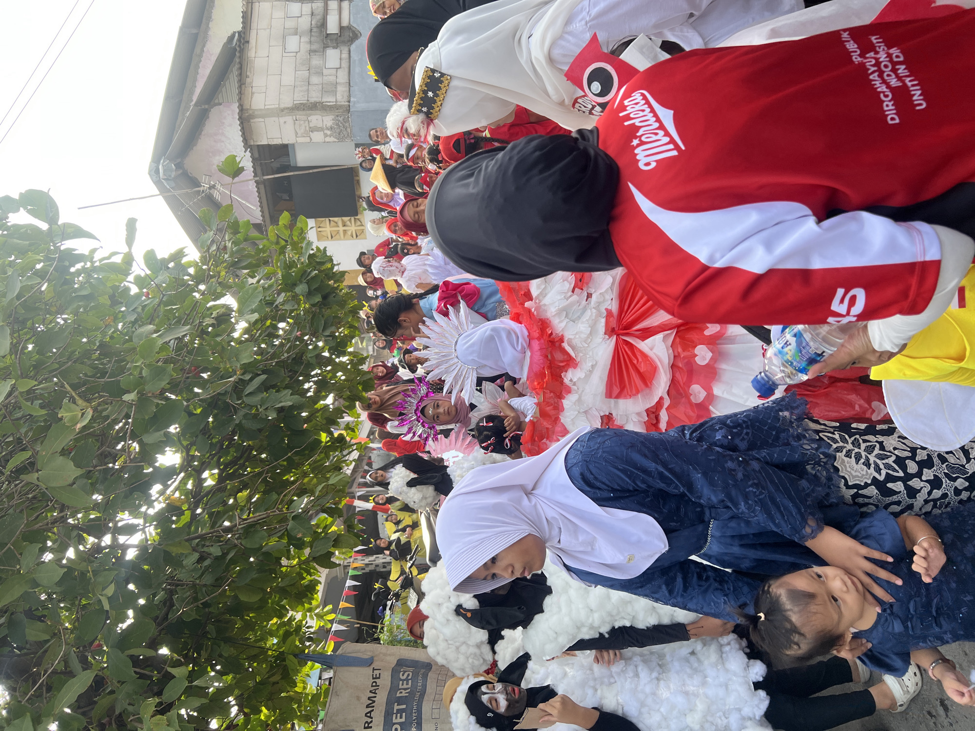 In Klapanunggal, Indonesia, a group of people, predominantly women and children, are participating in what appears to be a parade or festival. The participants are dressed in various costumes. Some individuals are adorned in outfits that resemble sheep, covered in white fluffy material and with black face paint. Others are wearing elaborate costumes, including a large red and white dress with intricate paper decorations. Many participants are wearing hijabs, with colors ranging from white and blue to black. One person in the foreground wears a red shirt with white detailing and the word "Merdeka" printed on it, suggesting a celebration of Indonesian independence. Another individual in the background is wearing a yellow conical hat. The environment shows a street scene with buildings visible in the background, including a structure with a corrugated metal roof. Trees with green foliage are present overhead, and decorative flags are strung across the area. A plastic water bottle is held by one person, and a motorcycle is parked behind some of the participants. The overall atmosphere appears festive and communal.