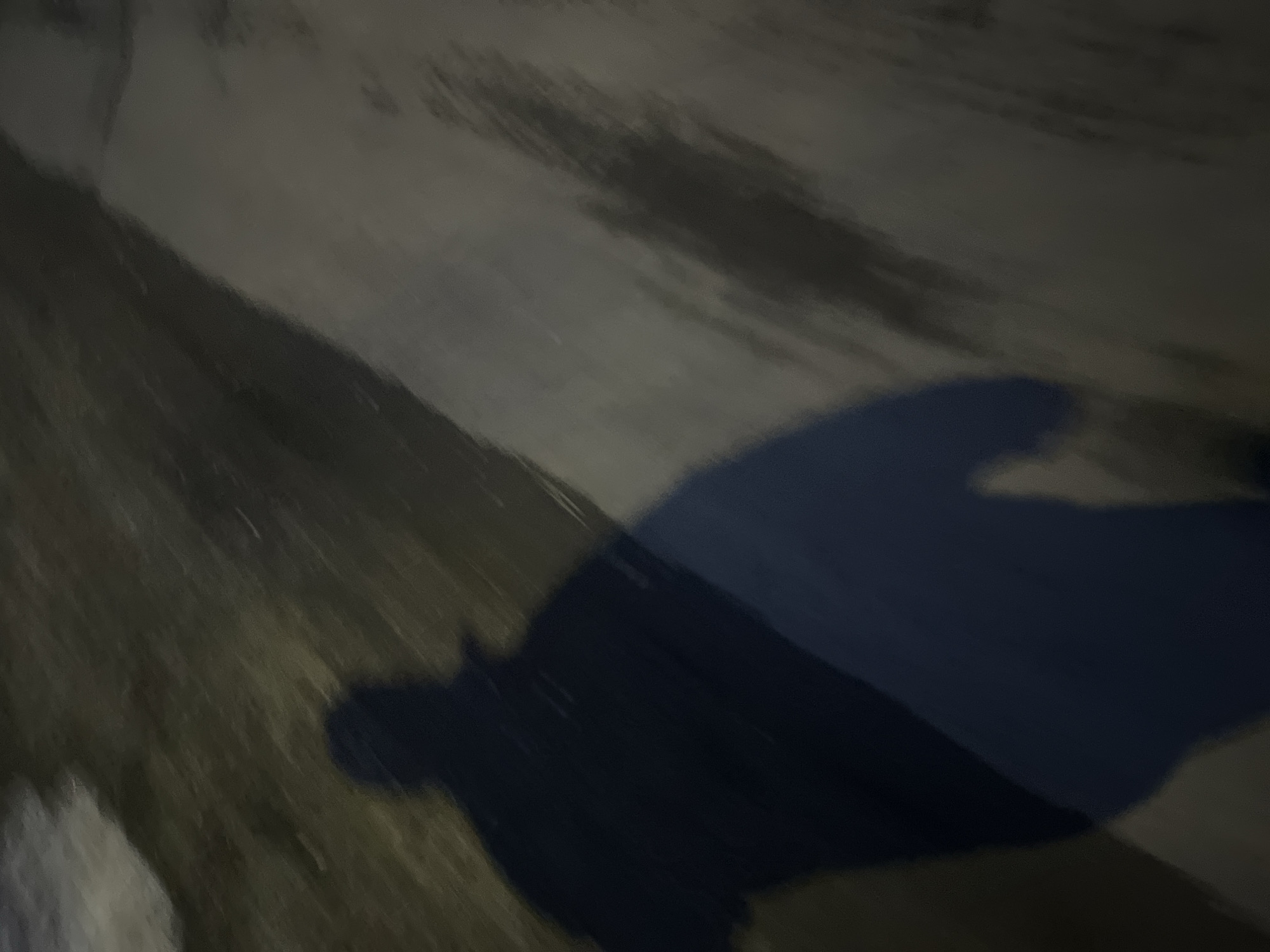 The image shows a blurry, low-light scene. In the center is a dark, indistinct shadow that appears to be the shadow of a person. The background consists of uneven surfaces, possibly a sidewalk or ground covered in patches of grass and light-colored material. Due to the lack of clarity and details, a specific location in Podolsk, Russia cannot be determined.