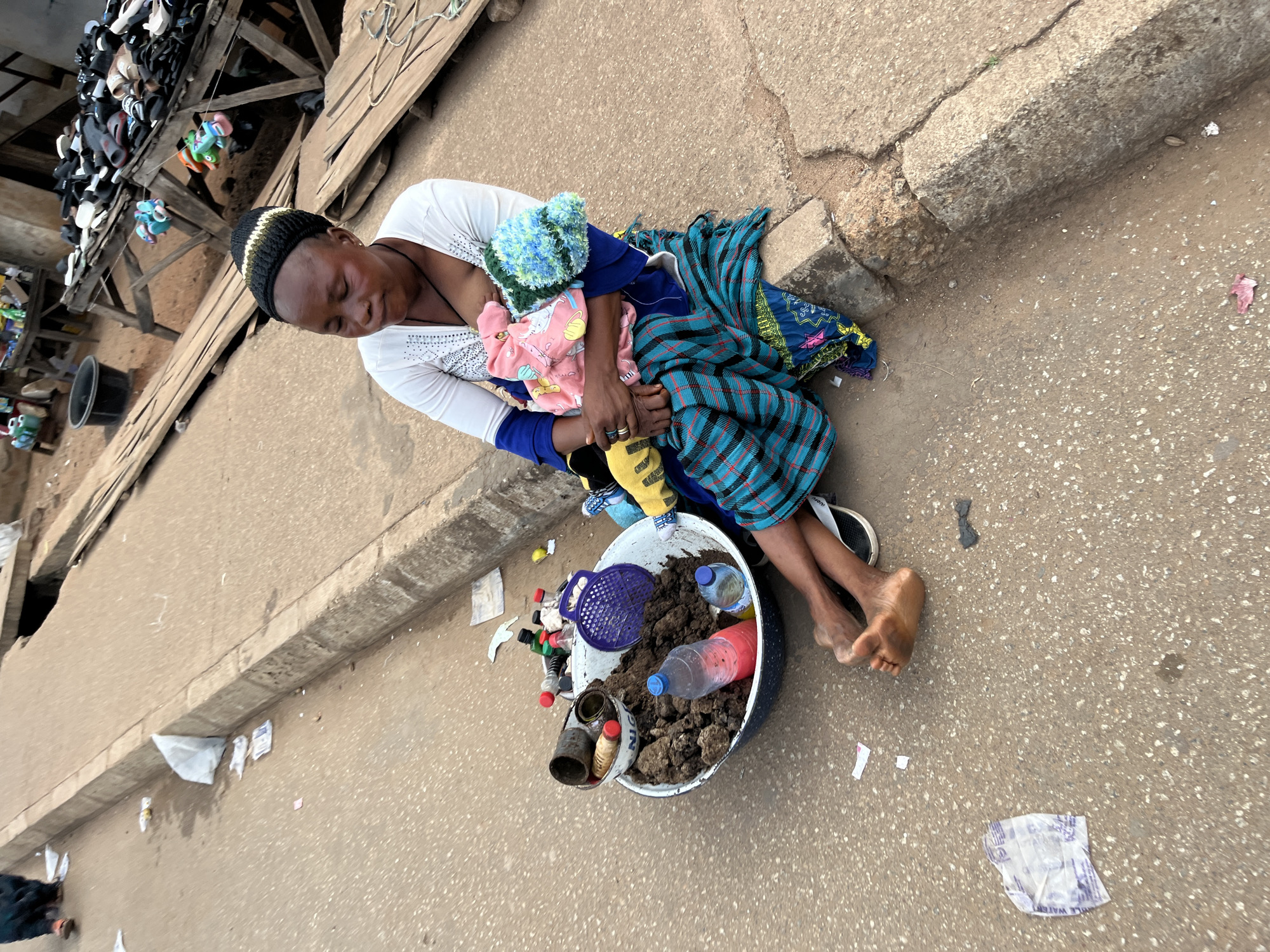 A woman is sitting on a paved sidewalk in a busy marketplace in Bukuru, Nigeria, nursing a baby. The woman is wearing a white long-sleeved shirt and a blue and green checkered skirt. She has a black and yellow knitted hat on her head. The baby is wrapped in a pink and yellow blanket and has a blue knitted hat on its head. The woman is holding the baby in her arms, and the baby is feeding from her breast.
In front of the woman is a large metal bowl filled with soil, several plastic bottles, and some tin cans. There is also a purple plastic strainer in the bowl. The sidewalk is littered with trash, including plastic bags, wrappers, and discarded items. In the background, there are market stalls with various goods for sale, including shoes and clothing. The overall scene suggests a bustling and somewhat unkempt urban environment.