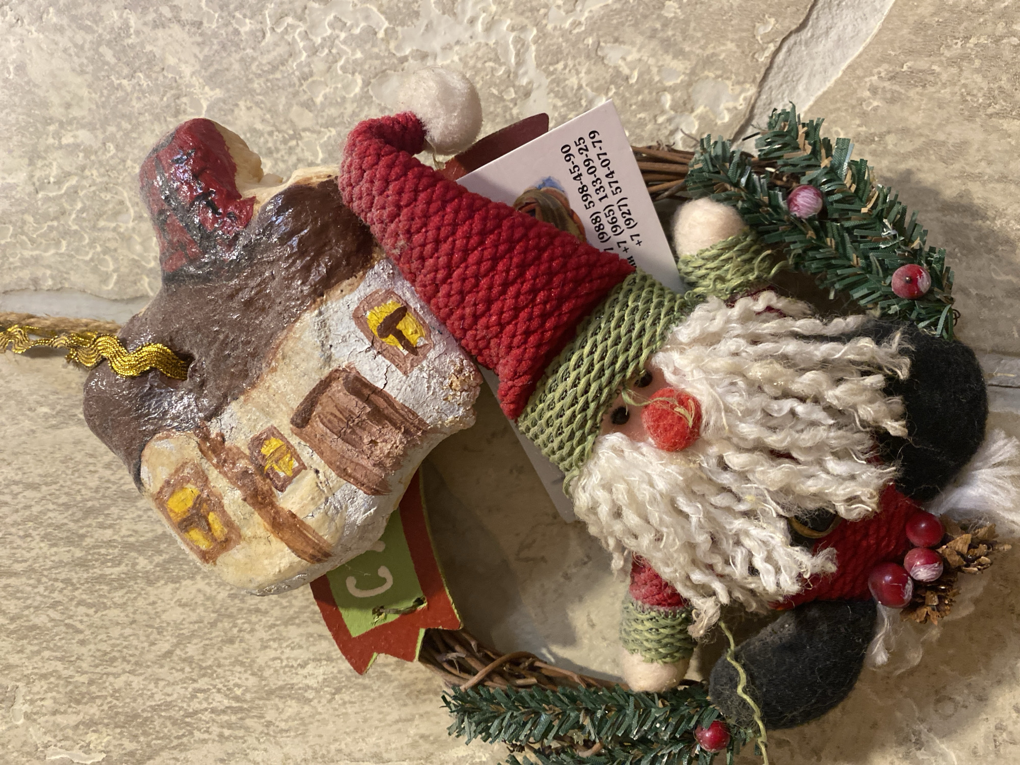 A close-up, top-down, outdoor shot shows a handmade Christmas ornament of Santa Claus and a house, hanging against a textured, light brown wall. The ornament is made of fabric and wood, with a red hat, green scarf, and white beard on Santa. The house is brown with yellow windows. A small tag with Russian phone numbers is attached to the ornament.