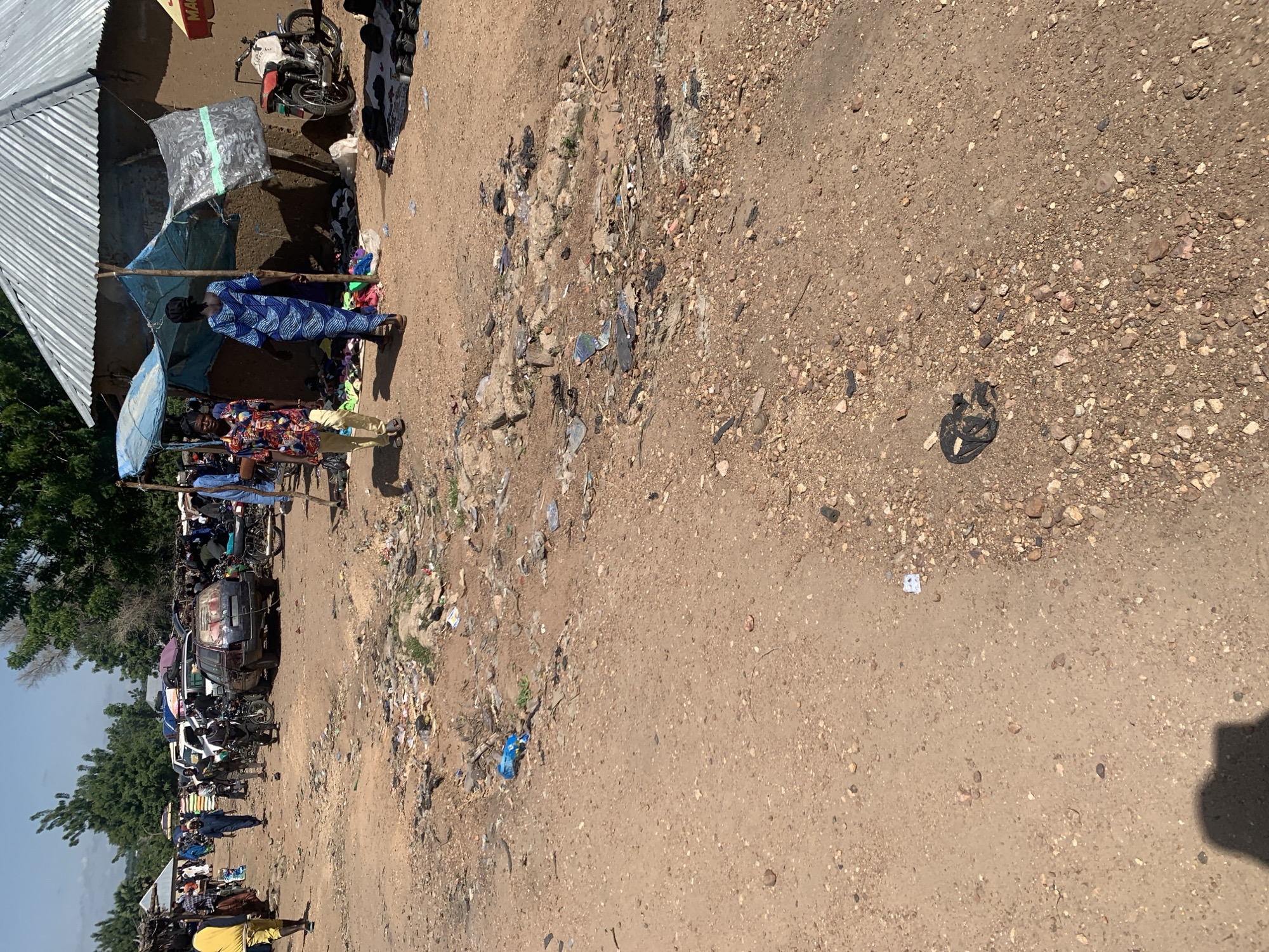This is a ground-level, outdoor, medium shot of a bustling outdoor market in Dull A, Nigeria. The scene is chaotic and dusty, with a mixture of people, vehicles, and merchandise. A dirt path runs through the middle of the frame, littered with debris and trash.
In the foreground, a person is walking from right to left, wearing a blue and white patterned dress and a headscarf. To their left, another person is walking from right to left, wearing a brightly colored short-sleeved shirt and light-colored pants. Behind them, several other people are visible, some walking and some standing.
Several vehicles are present in the midground, including cars, motorcycles, and what appears to be a truck. Some of the vehicles have luggage strapped to their roofs. Tents and makeshift shelters are set up, providing shade for vendors and shoppers. Merchandise, including clothing and other items, is displayed on the ground and under the shelters.
The background is filled with trees and buildings, suggesting a densely populated area. The sky is clear and blue, indicating a sunny day. The overall impression is one of a lively, but somewhat disorganized, marketplace.