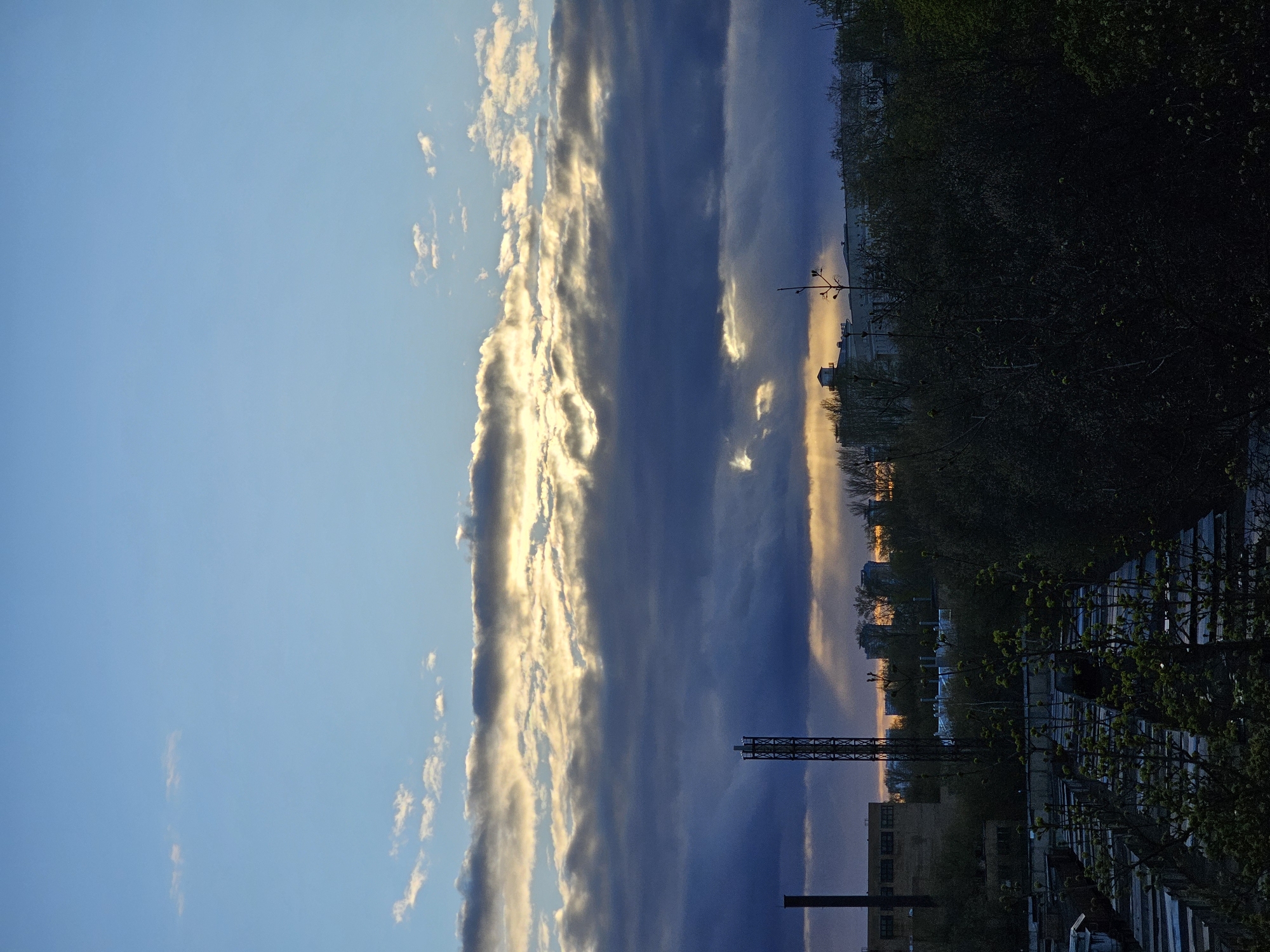 The image captures a dramatic sunset or twilight over an urban-industrial landscape in Khimki, Russia. The sky is the dominant feature, presenting a striking contrast. On the left, it transitions from a clear, light blue with scattered wispy white clouds. Towards the center and right, a dense, horizontal band of clouds dominates, their undersides a deep grey and blue, while their upper edges and sections illuminated by the setting sun glow intensely with golden-white light. The sky beneath these clouds deepens to an indigo and purplish hue.
Along the distant horizon, a bright golden-orange glow indicates the position of the setting sun, visible behind a silhouette of distant buildings and a continuous line of dark trees and foliage.
In the foreground, various structures and foliage are rendered in dark silhouettes against the brighter sky. A prominent, tall metal lattice tower stands on the left. Below it, the upper portions of a dark, multi-story building and a vertical stack or chimney are visible. The right side of the foreground is heavily wooded with dense, dark trees, some displaying emerging green leaves, suggesting late spring or early summer.
No people or specific activities are discernible. The scene emphasizes the atmospheric quality of dusk, with the interplay of intense light and shadow from the setting sun creating a serene yet dramatic ambiance over a developed area.
