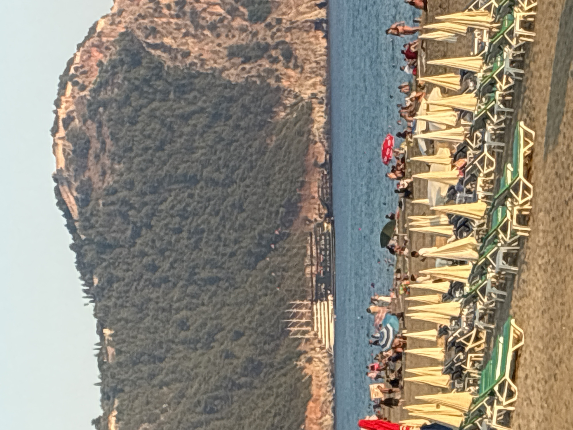 The image depicts a crowded beach in Alanya, Turkey, with the Alanya Castle ruins prominently visible on a mountain overlooking the scene. The beach is lined with rows of empty lounge chairs and beach umbrellas, most of which are a pale yellow color, suggesting it is a popular tourist destination. In the foreground, several green and white lounge chairs are occupied by sunbathers. The sea is a calm blue, with a large, multi-masted ship sailing in the distance. The mountain behind the ship is covered in dense green foliage, with the ancient stone structures of the castle perched on its summit, catching the warm light of what appears to be late afternoon. The sky is a clear, pale blue. The overall impression is one of a vibrant and bustling beach resort.