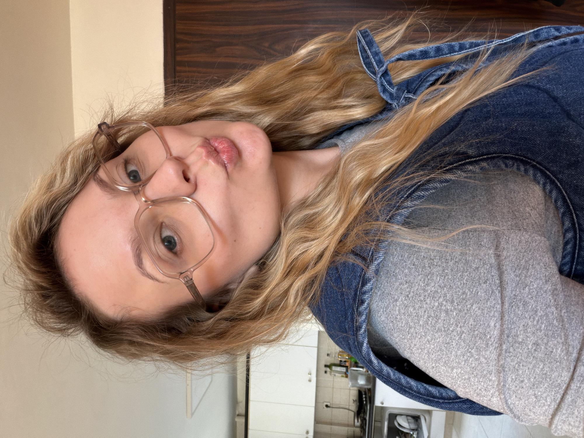 A woman with long blonde hair and glasses is posing for a selfie. She is wearing a denim jacket over a gray shirt and has her lips pursed. Behind her is a kitchen with white cabinets and appliances. The location of the city of Tel-Aviv, Israel, cannot be determined from the image.