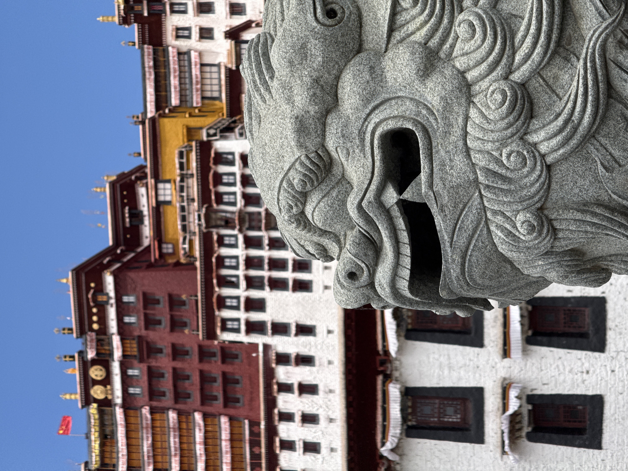 A stone lion head statue is in the foreground, appearing to be mounted to a wall or ledge of the Potala Palace in Chengguan District, Lhasa, Tibet Autonomous Region, China. The Potala Palace, a large complex of buildings with white, red, and gold facades, is partially visible in the background under a blue sky. The buildings exhibit a pattern of multiple dark-framed windows. A red flag is seen on one of the gold-adorned roofs of the palace.