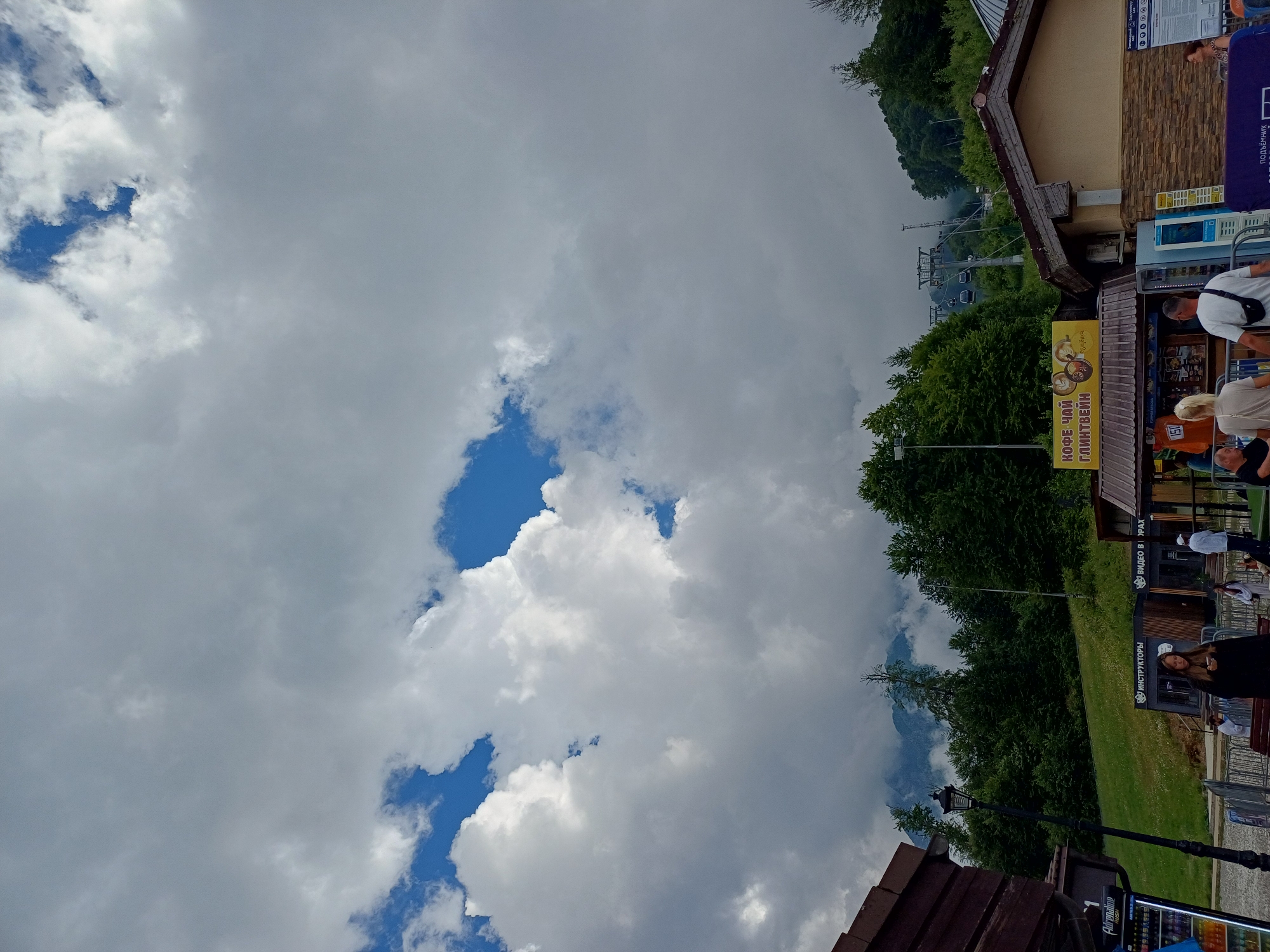 The image depicts a view in Estosadok, Russia, with a backdrop of lush green trees and mountains shrouded in clouds. In the foreground, there are several structures, including a wooden building on the left and a building with a stone facade on the right. Between these buildings, there are food kiosks with signs in Russian advertising coffee, tea, and mulled wine. People are seen in the foreground, some interacting with the kiosks and others walking. A ski lift with several cabins can be seen ascending the mountainside in the distance. The sky is filled with dramatic, large cumulus clouds, with patches of blue sky visible through the gaps. The overall atmosphere suggests a resort area, possibly during a cloudy summer day.