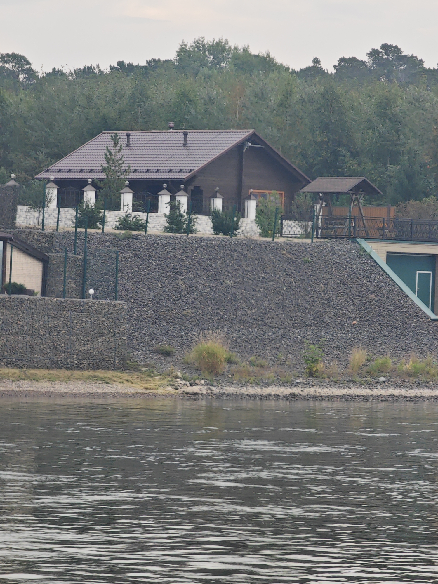 The image shows a large, dark wooden house with a brown shingled roof, situated on a steep, rocky embankment. The house has several windows with dark frames and appears to be two stories high. In front of the house, there is a white stone fence with decorative posts topped with lights. Several small trees and bushes are planted along the fence line.
To the right of the house, there is a dark wooden gazebo with a brown roof, and beyond that, a wooden fence. A green structure with a white-framed door is visible further down the embankment to the right, possibly a garage or storage unit. The embankment itself is covered in dark gray stones and small patches of dry grass and vegetation.
Below the embankment is a narrow strip of rocky shore, and then a body of water, likely a river, with visible ripples and reflections. The background is dominated by a dense forest of green trees under a cloudy sky. The overall scene suggests a rural or semi-rural residential setting. There are no people visible in the image. The location is identified as Ермолаево, Russia.