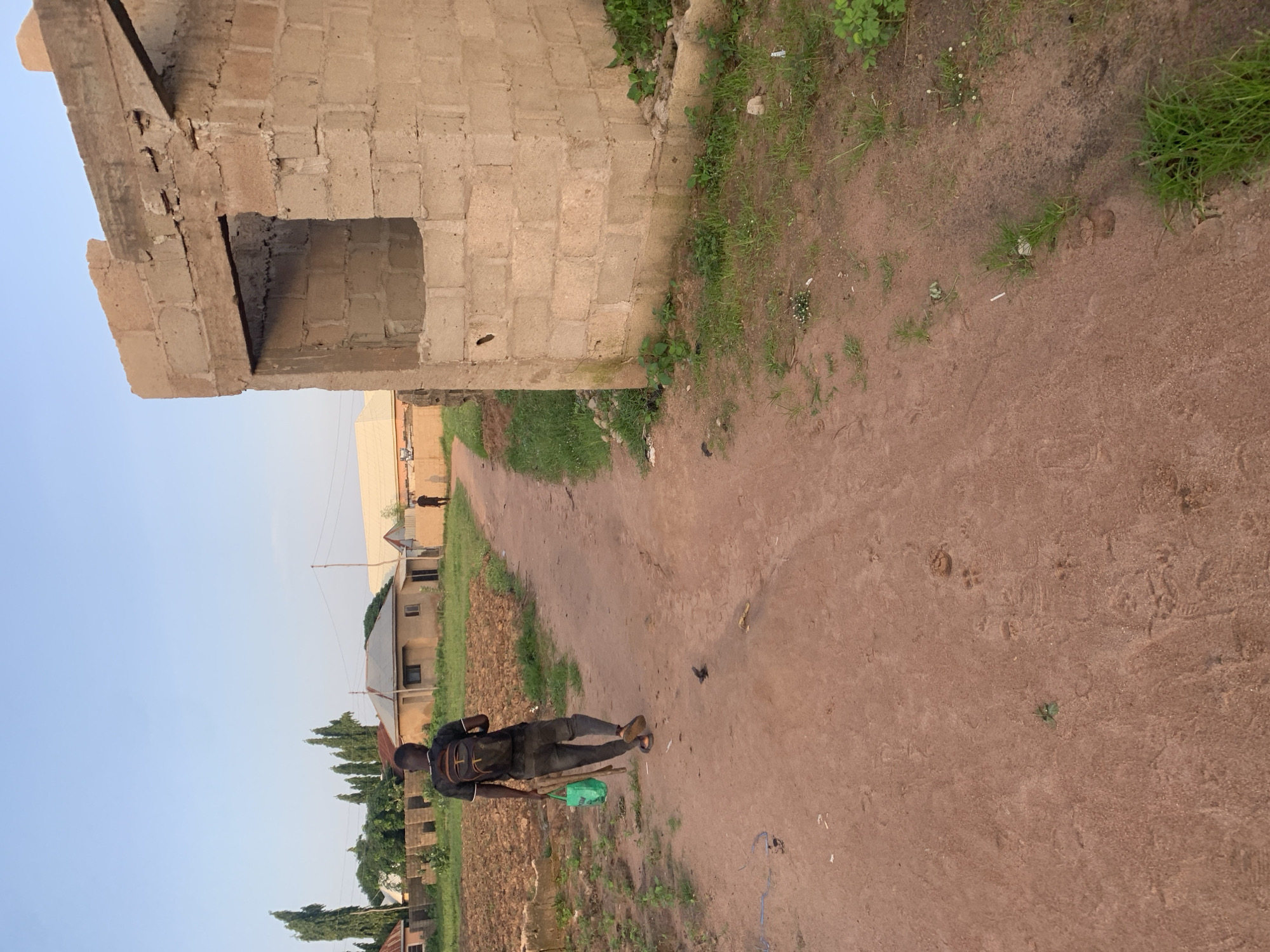 A young man wearing a black t-shirt, black pants, and a backpack walks away from the camera on a dirt road. He carries a green bag in his left hand. He is in an unpaved area, likely in Jalingo, Nigeria, with several houses visible in the background, some appearing to be under construction. The road is flanked by a cultivated field on the left and a partially constructed brick building on the right. The sky is clear and blue, suggesting it is daytime.