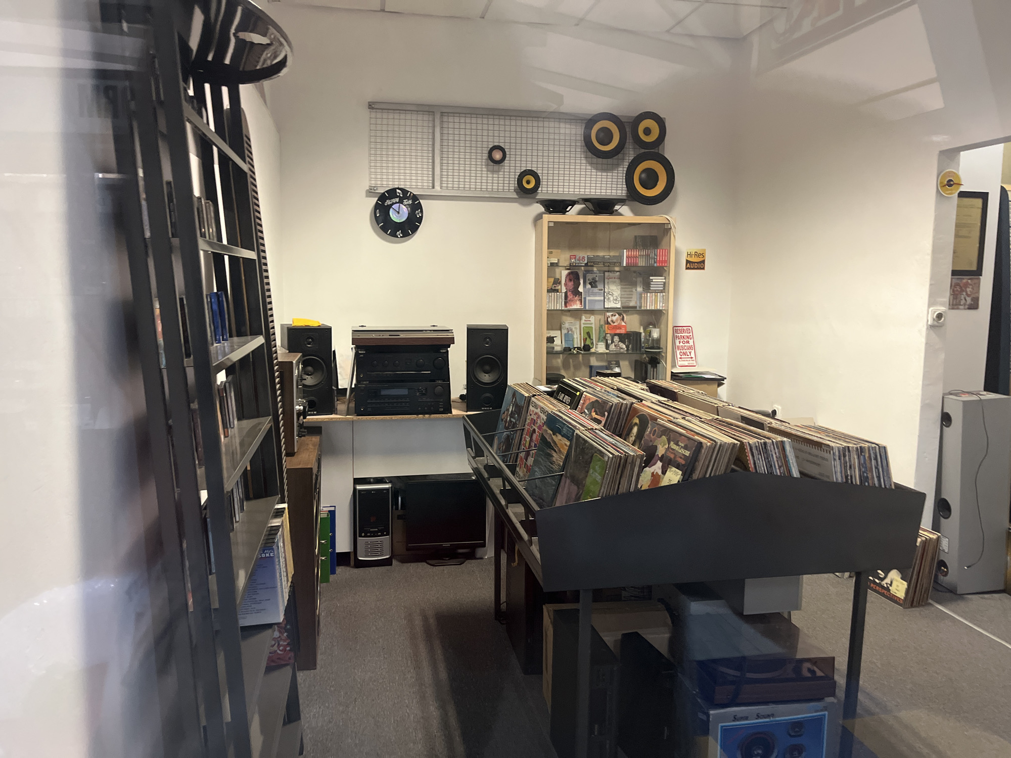 This image depicts the interior of what appears to be a record store or an audio equipment shop, likely located in Plovdiv, Bulgaria. The room has white walls and a gray carpeted floor, with notable reflections in the foreground suggesting the photo was taken through glass.
In the left foreground, a tall, dark, multi-shelved bookshelf is visible. Towards the center, an audio setup is arranged on a shelf, featuring two black bookshelf speakers, a stack of audio components (including a receiver and possibly a turntable or amplifier), some with wood-grain detailing. Below this, a silver desktop computer tower and a flat-screen monitor are placed on the floor.
The central wall is decorated with a large metal grid panel. Mounted on this grid are several speaker cones of various sizes in yellow-and-black, along with one small pinkish-brown cone, arranged decoratively. To the left of the grid, a black wall clock designed like a vinyl record displays the partial text "OKE."
To the right, a wooden display cabinet with glass doors showcases a collection of CDs and cassette tapes, one of which shows the number "46." A yellow-and-black sign on the cabinet reads "Hi-Res AUDIO," and below it, a red-and-white sign states "RESERVED PARKING FOR MUSICIANS ONLY."
The mid-ground is dominated by a large, black metal display rack filled with numerous vinyl records, presented upright for browsing. Visible album titles on the covers include "EARL HINES" and "PERSPECTIV," with "Sunce Scaune" also visible on a record in the lower right. A tall, slender gray speaker column stands against the wall on the far right. No people are visible, focusing attention on the merchandise and decor.