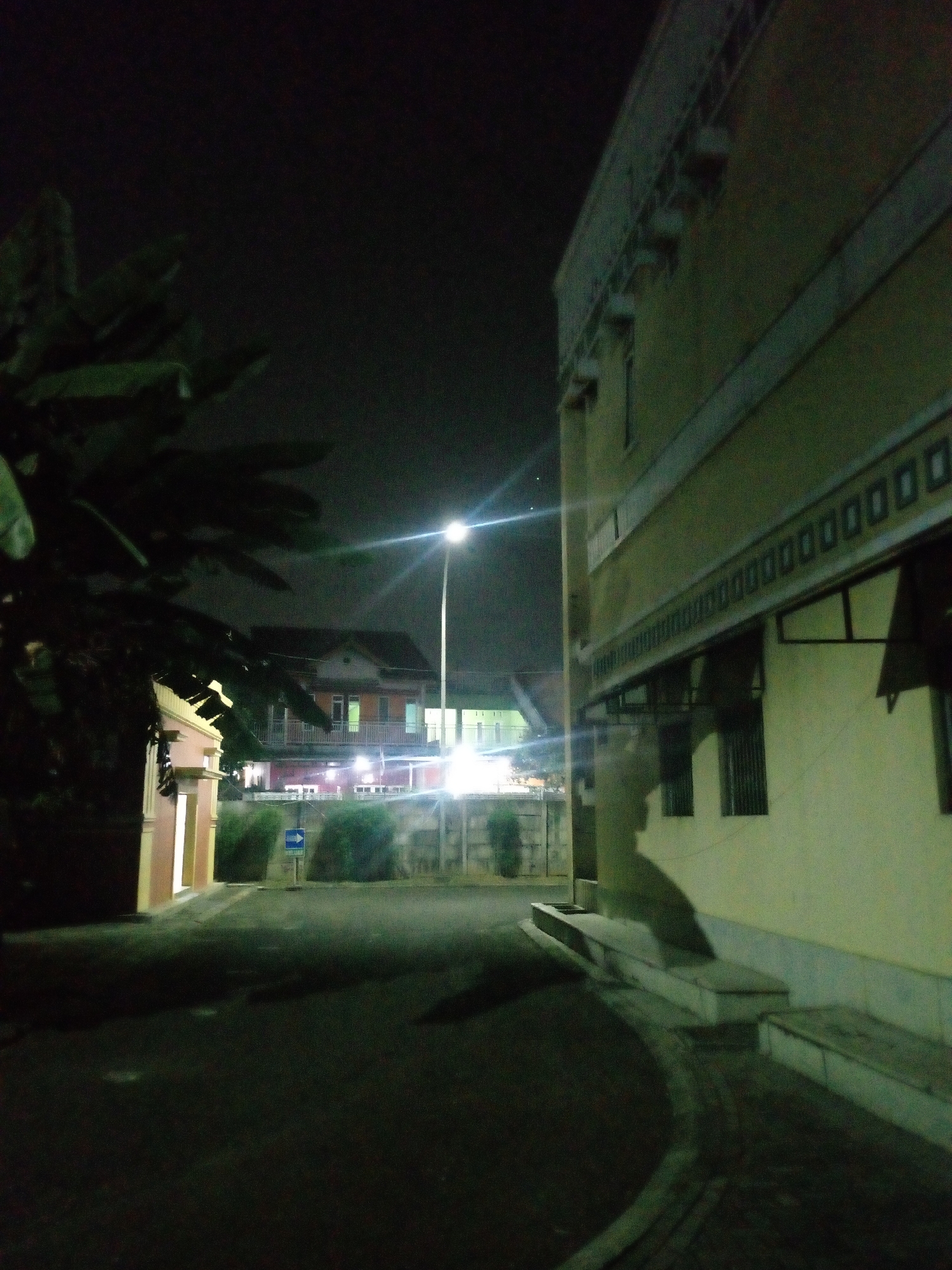 A night scene unfolds on a paved road, possibly within a residential or institutional complex in Bitung Jaya, Indonesia. A prominent, pale yellow building with dark-framed windows and decorative molding runs along the right side of the frame, its facade illuminated by artificial light. Opposite this building, to the left, a lighter-colored structure with a pinkish hue is partially visible, with large green leaves from a tree casting shadows across its surface.
In the mid-ground, a street lamp emits a bright light, casting a glow on the surrounding area and creating lens flare. Behind it, a multi-story building with orange and white elements is visible, its windows lit from within. A blue directional sign indicating a right turn is posted near some dense green bushes, situated between the two main structures. The road surface appears dark and wet in places, reflecting the ambient light. The sky above is black and devoid of stars, suggesting a cloudy or heavily light-polluted environment. The overall impression is one of a quiet, dimly lit urban or suburban setting at night.