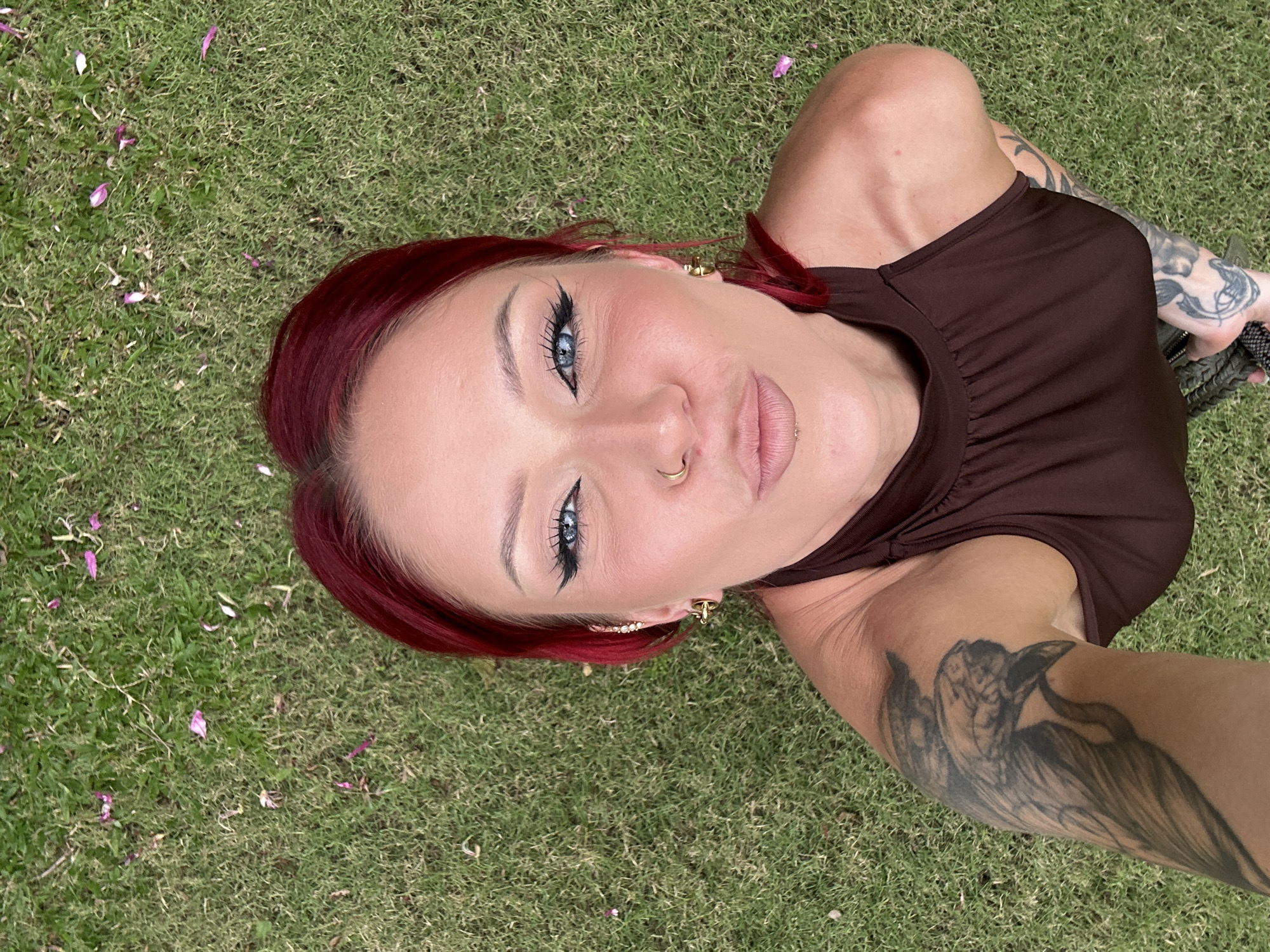 A woman with red hair and tattoos on her arms lies on a patch of green grass in Nusa Dua, Indonesia. She is wearing a brown sleeveless shirt and has a nose ring. Flower petals are scattered on the grass around her.