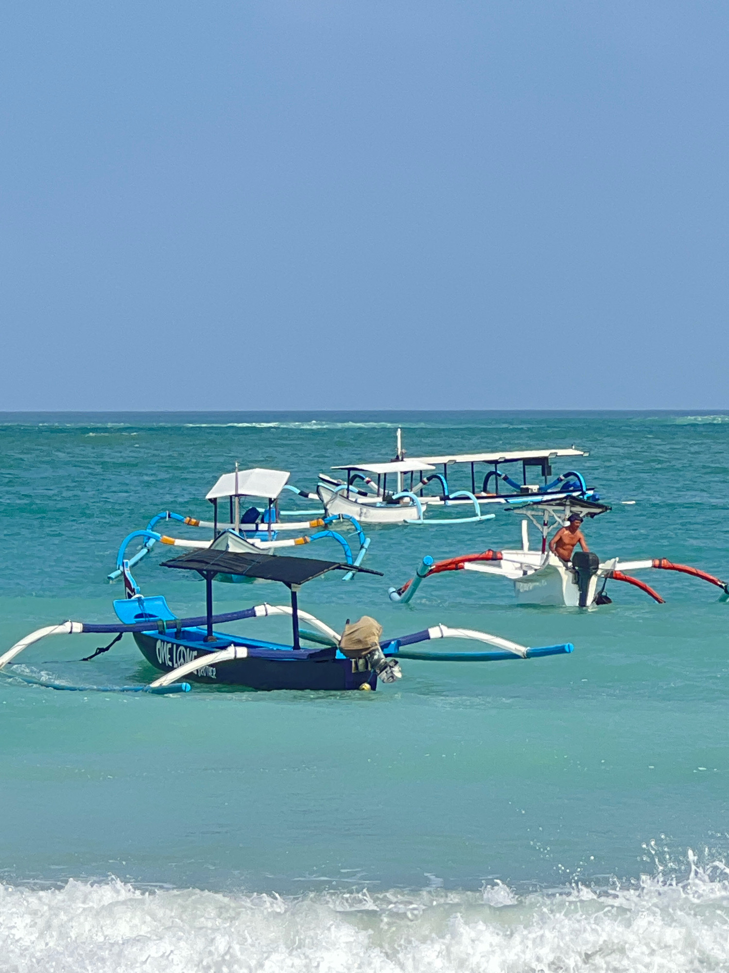 The image depicts a serene scene at the beach in Kuta, Indonesia, under a clear blue sky. Several traditional Indonesian outrigger boats, known as "Jukung," are anchored in the turquoise waters. In the foreground, a dark blue and white Jukung is prominently featured, with the words "ONE LOVE" visible on its side. A tan-colored canvas cover is draped over the back of this boat. Further out in the water, a white Jukung with red and white outriggers has a man with a tan complexion, wearing dark shorts, sitting in it. Another white Jukung, with a similar outrigger design and a white canvas canopy, is visible behind the first white boat. The gentle waves of the ocean create a tranquil atmosphere, characteristic of a beautiful day in Kuta.